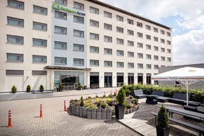 Un hotel con fachada moderna, entrada central y un patio con mobiliario y jardineras.