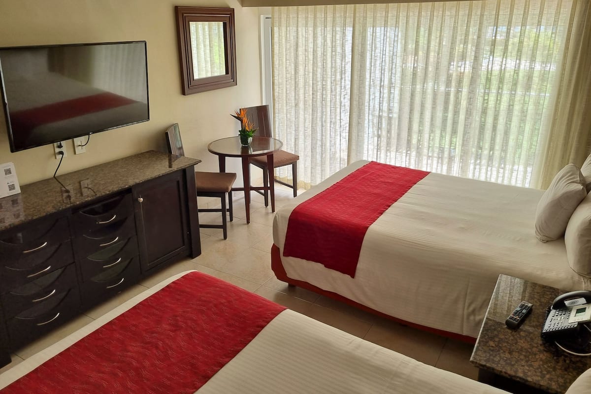 A hotel room with a bed, a desk with a telephone, a television, a small table with a chair, and a window with curtains.