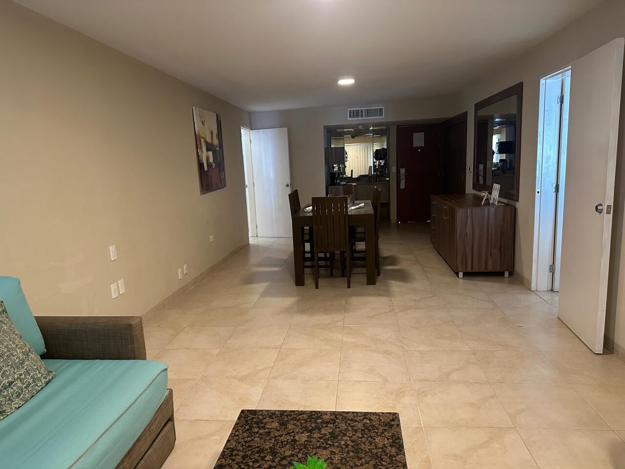 A living room with a sofa, a coffee table, a dining table with chairs, a kitchen area, and a hallway leading to a door.