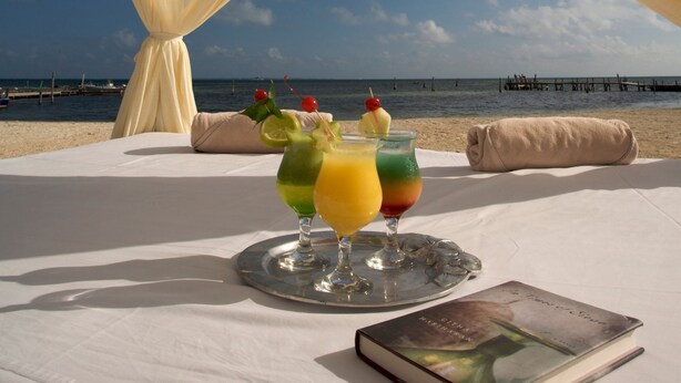 A beachside table with a drink and a book, set against a backdrop of a clear sky and ocean. - Ocean Spa Hotel – All Inclusive (Cancun)