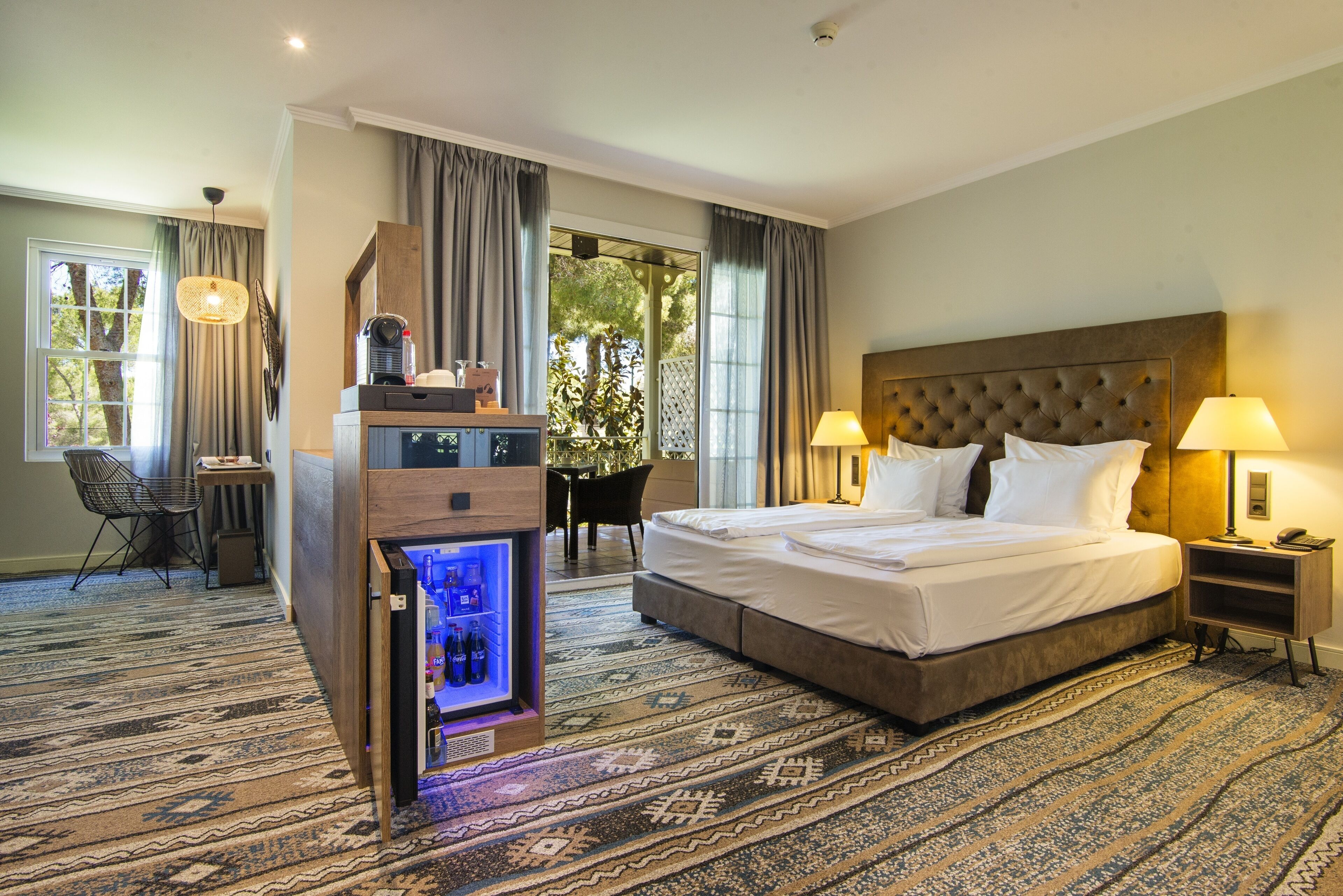 A hotel room with a large bed, a minibar, a desk with a chair, and a view of greenery through the window.