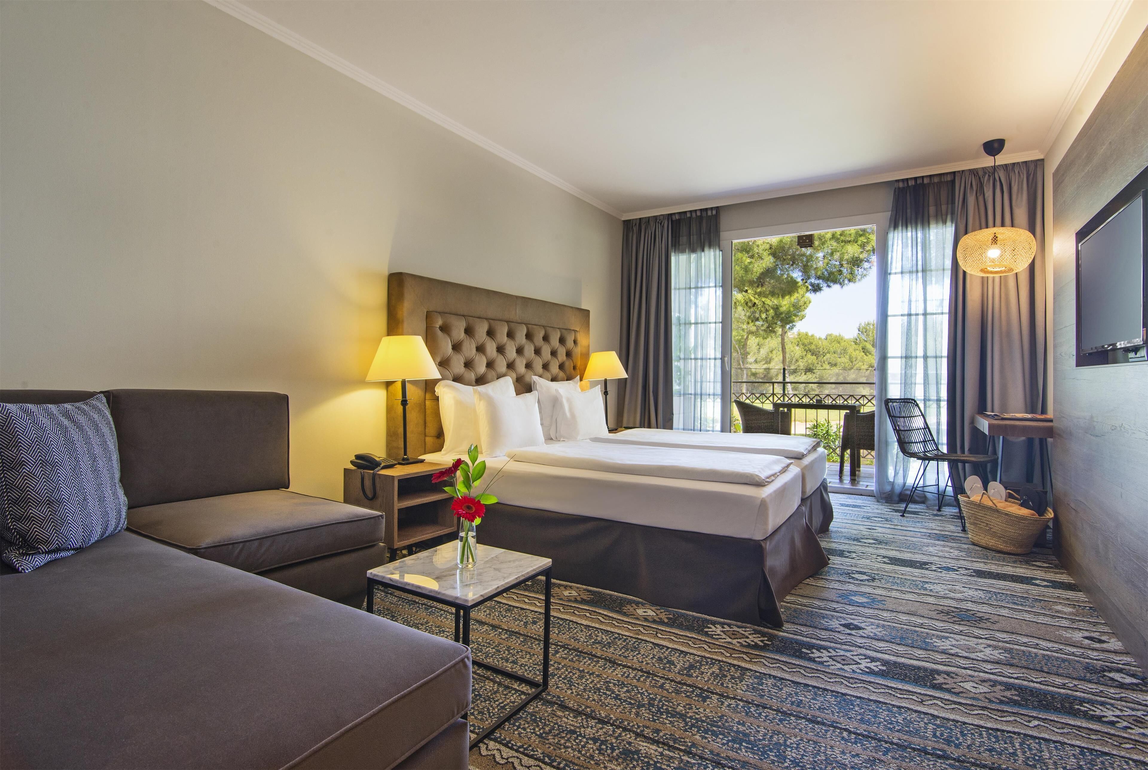 A hotel room with a large bed, a sofa, a small table with a vase of flowers, a TV, and a balcony with a view of trees.