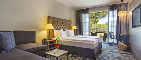 A hotel room with a large bed, a sofa, a small table with a vase of flowers, a TV, and a balcony with a view of trees.