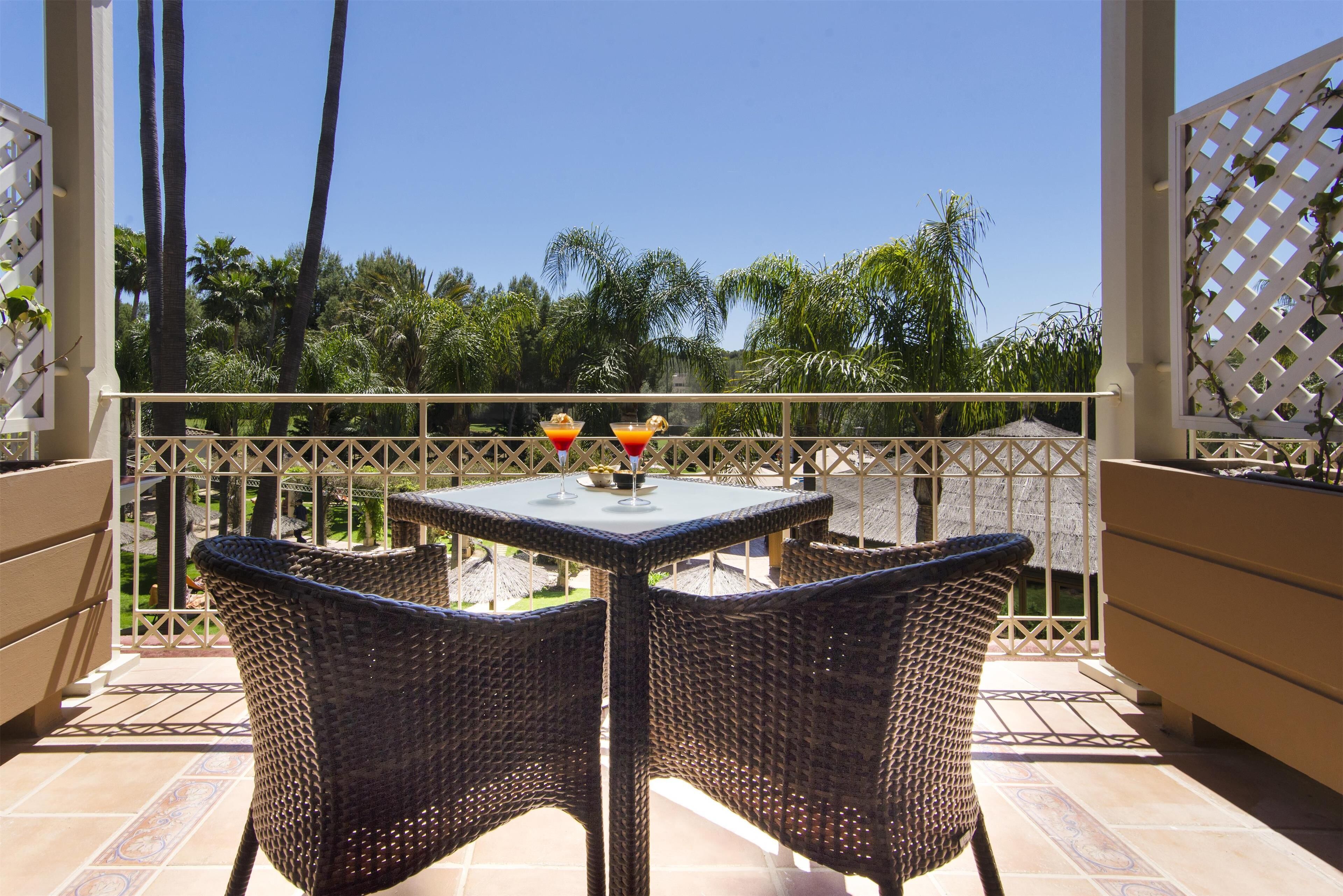 Un balcon avec des fauteuils en osier, une table avec un plateau en verre et une vue sur des palmiers.