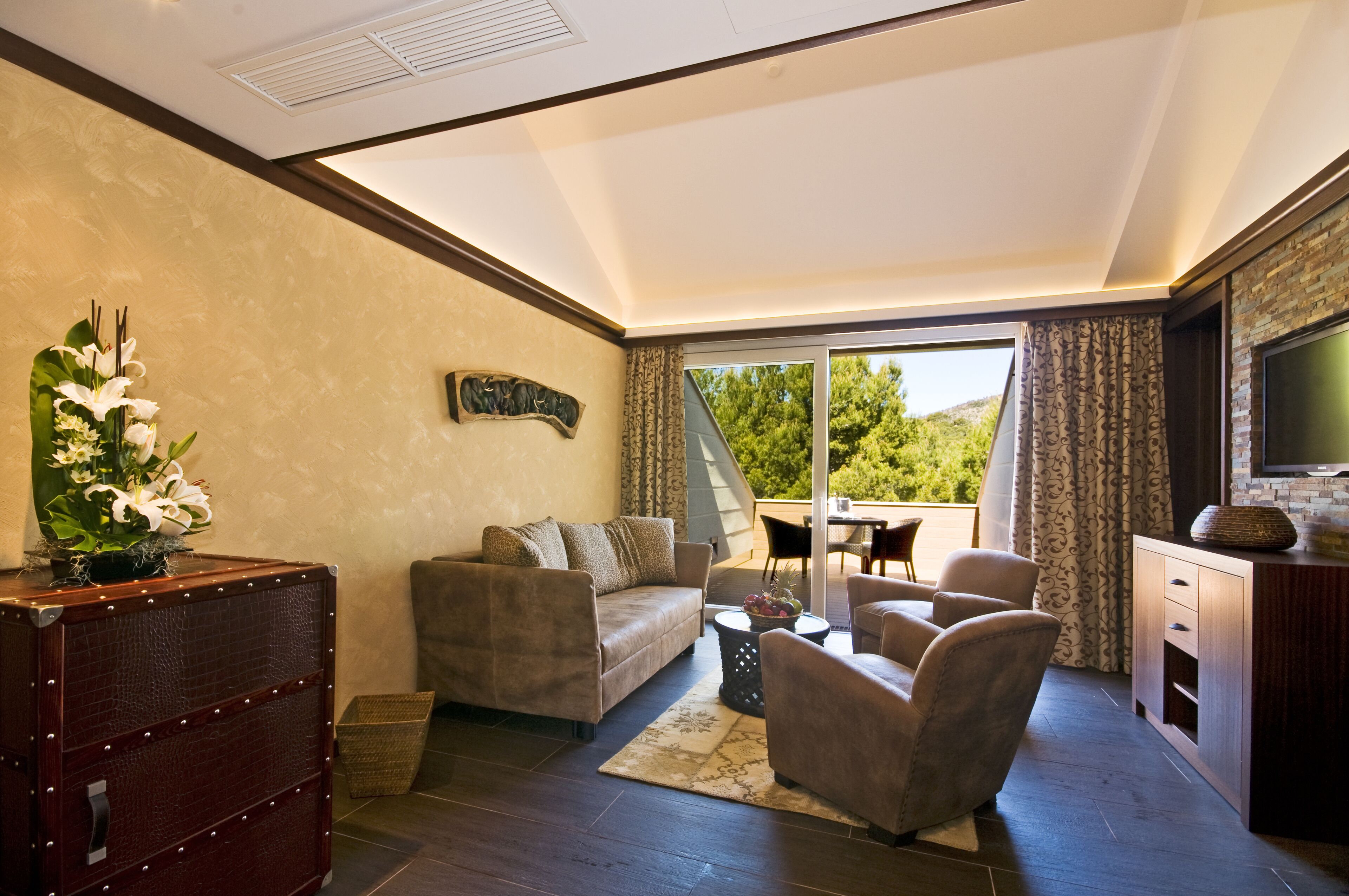 A modern living room with a sofa, armchairs, a coffee table, and a TV. There is a decorative vase with flowers, a wooden cabinet, and a large window offering a view of greenery.
