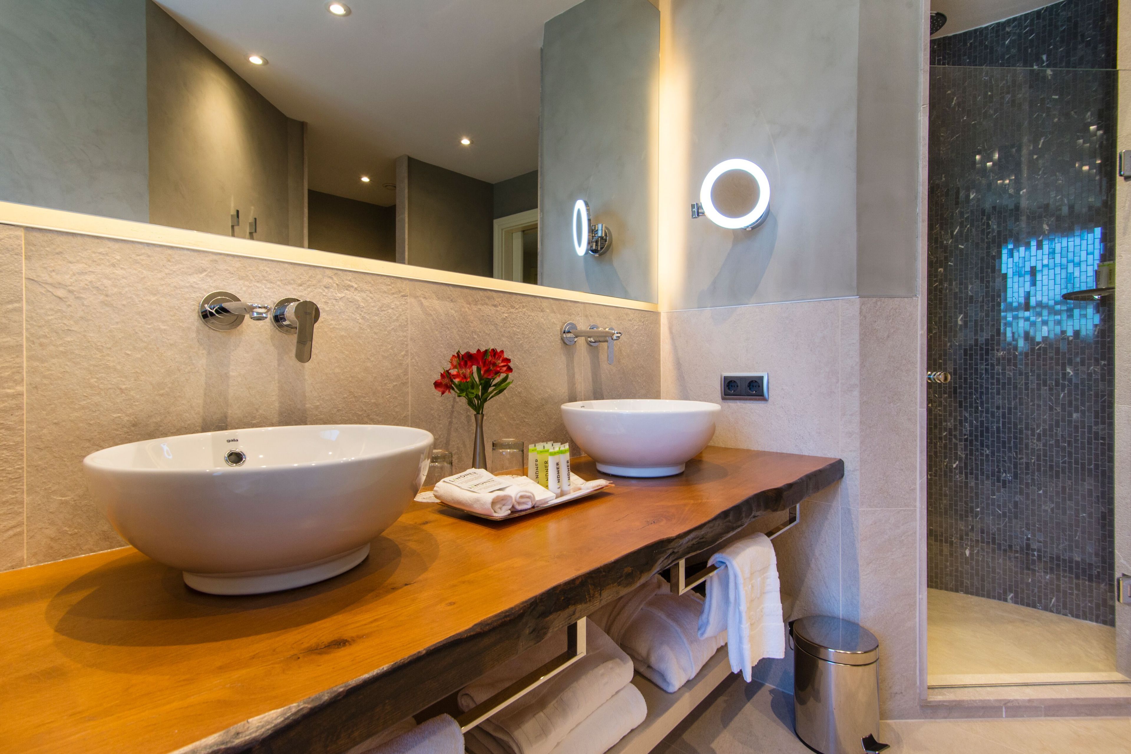 Un bagno moderno con due ampi lavandini, un mobile vanità in legno, uno specchio con illuminazione e una zona doccia.