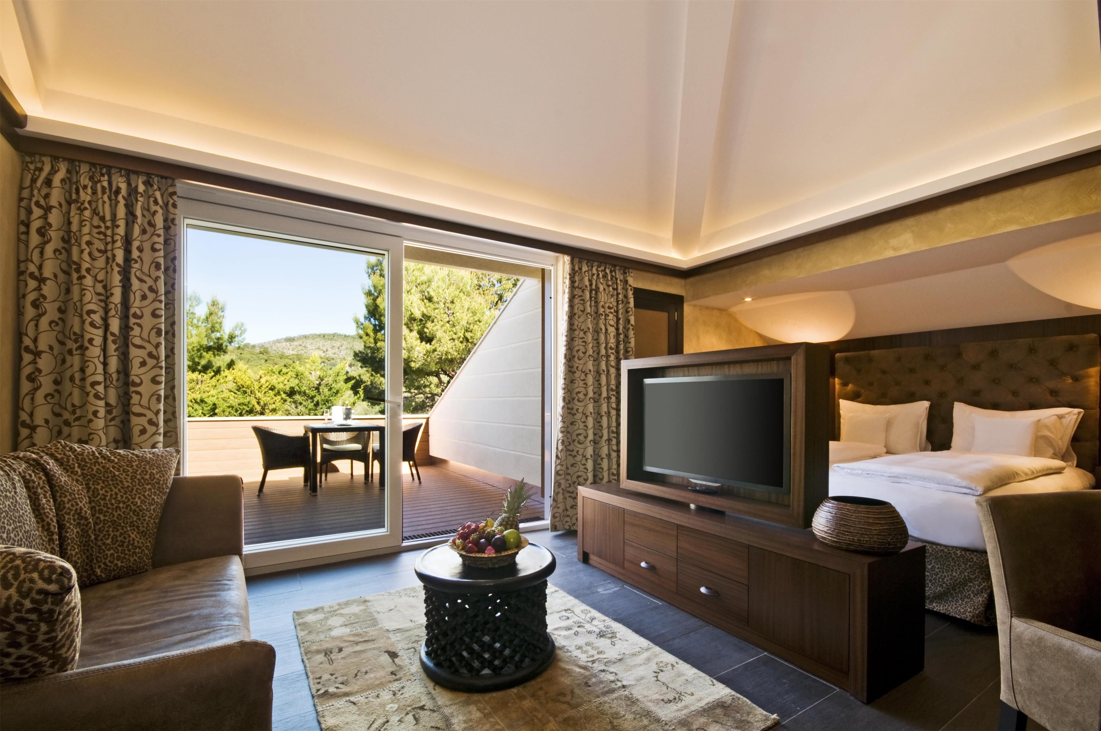 Habitación de hotel moderna con una cama grande, televisión, sofá y zona de comedor con vistas al exterior.