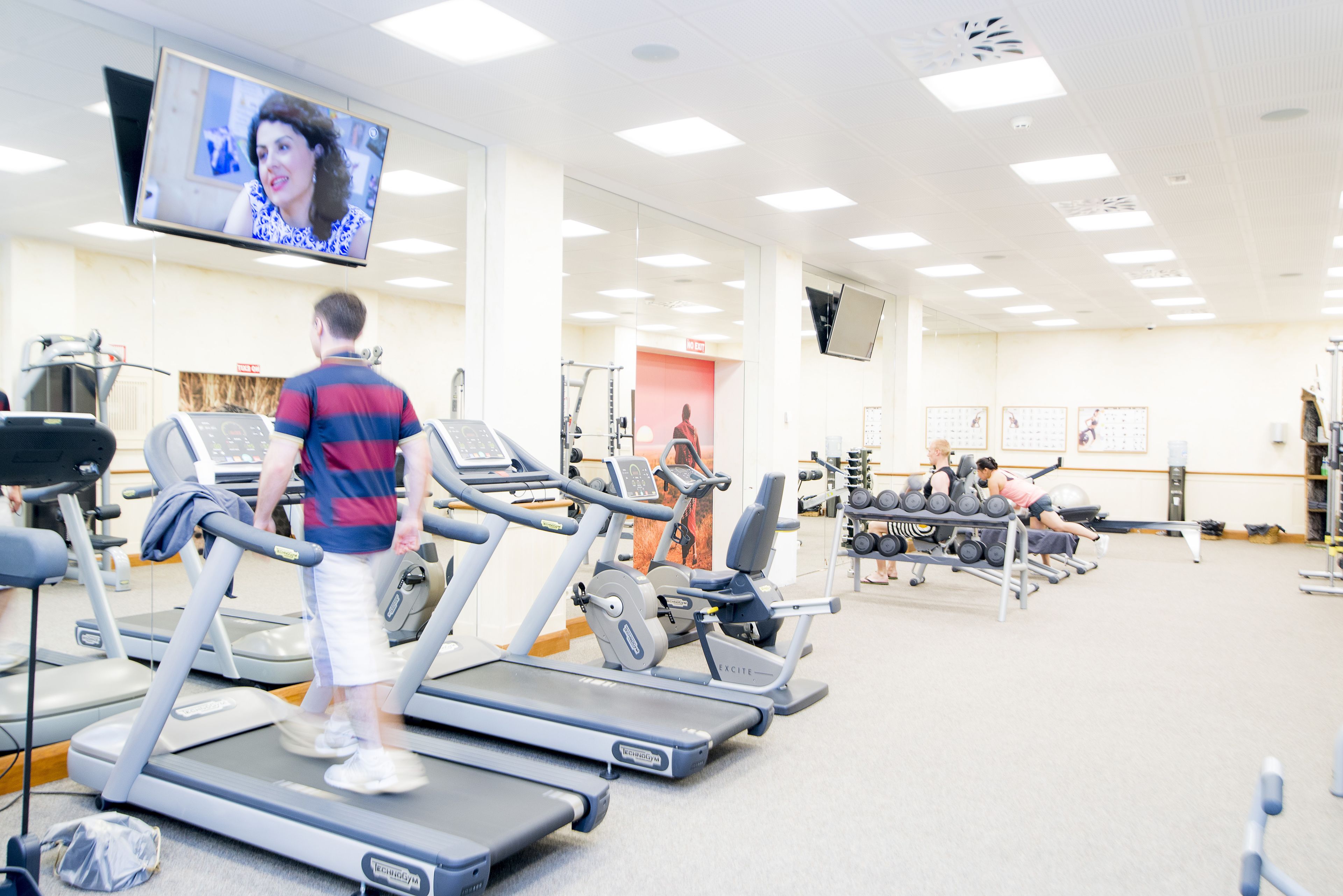 Ein Fitnessstudio mit Laufbändern, Kraftgeräten und einem Fernseher, auf dem eine Person zu sehen ist.