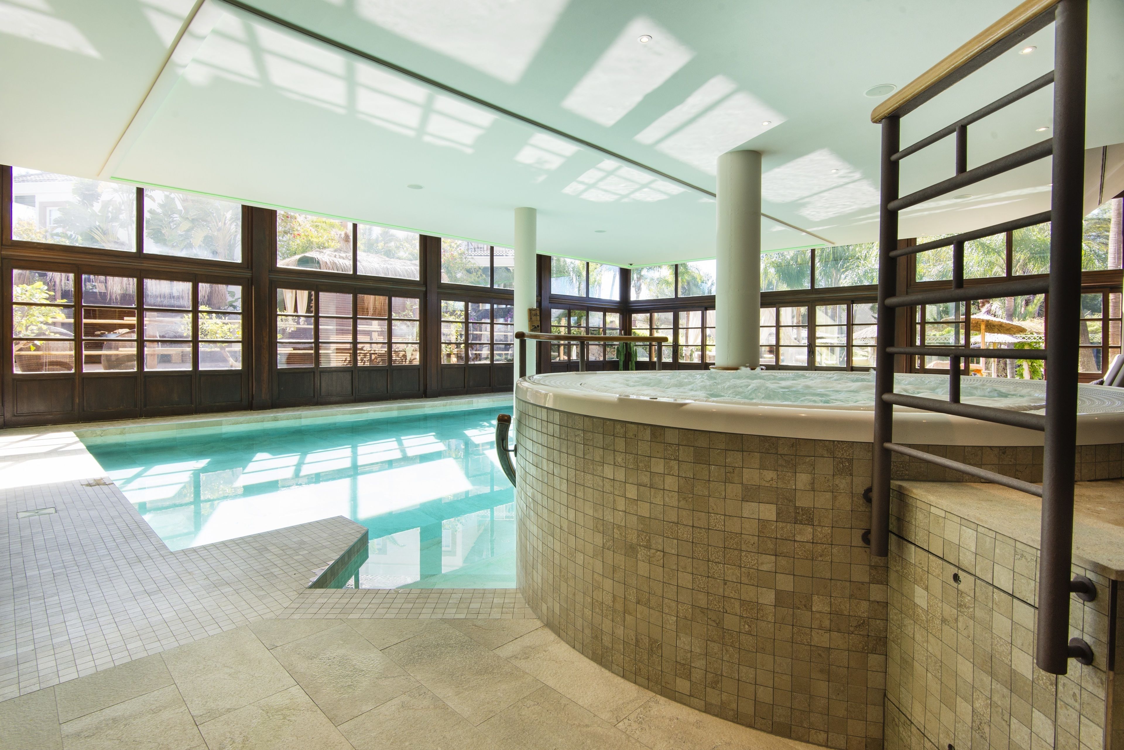 An indoor swimming pool with a hot tub, large windows, and a tiled floor.