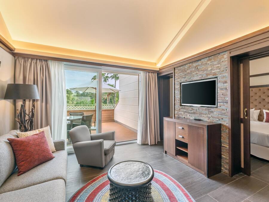 A modern hotel room with a sofa, armchair, TV, and a balcony with a table and chairs.