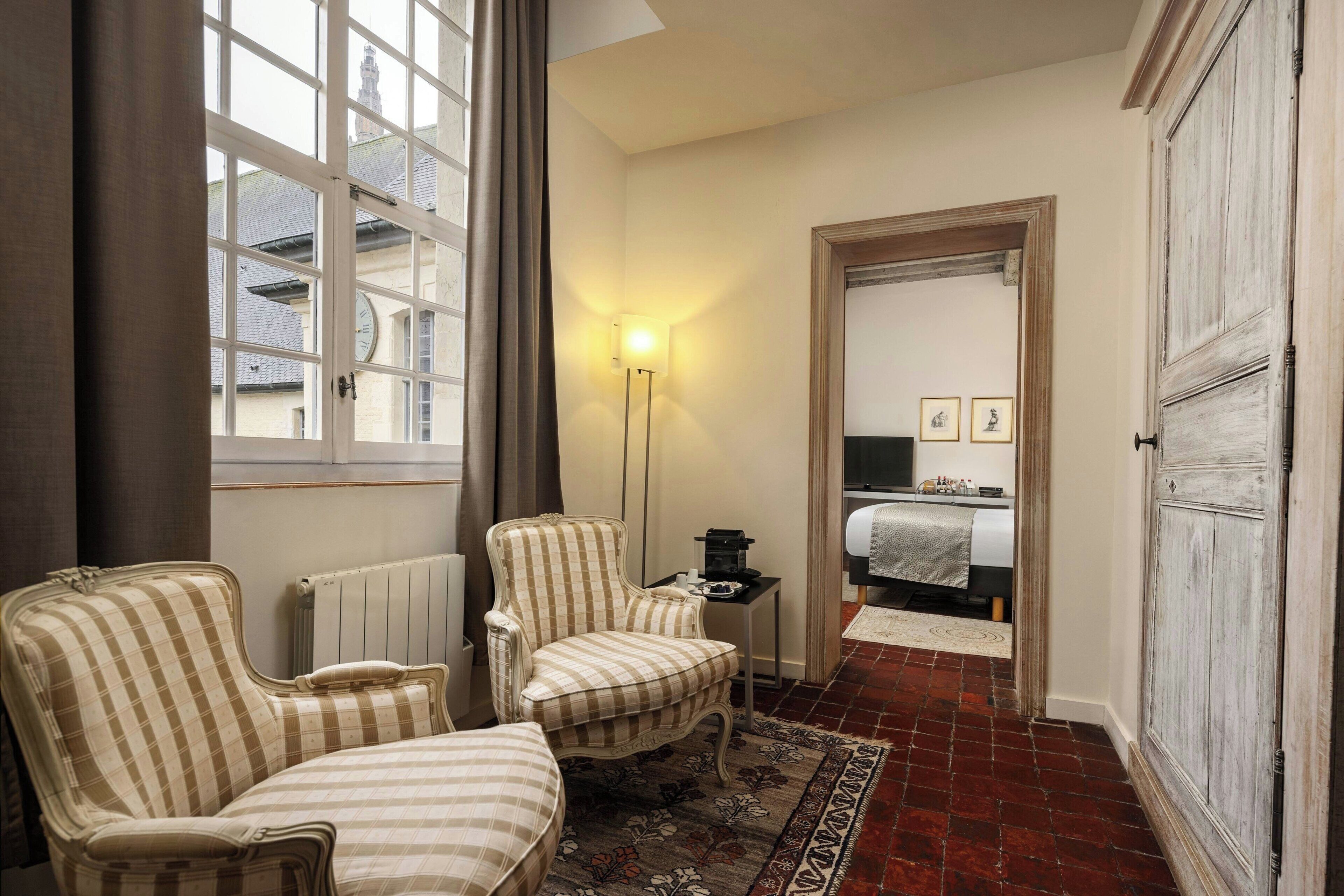 Una habitación con dos sillones a rayas, una mesita, un radiador, una lámpara de pie y vistas a un dormitorio a través de una puerta abierta.