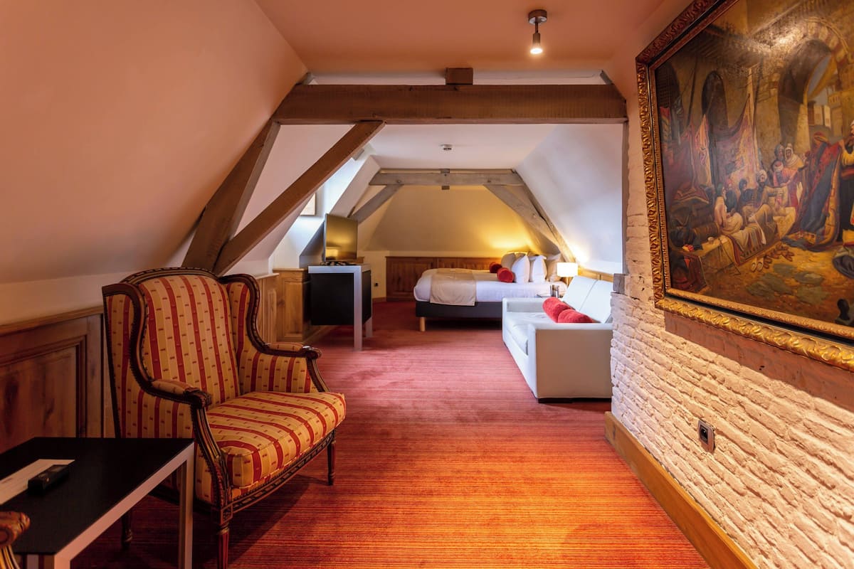 Une pièce avec un plafond en pente, un lit avec une literie blanche, une chaise, un bureau et un tableau accroché au mur.