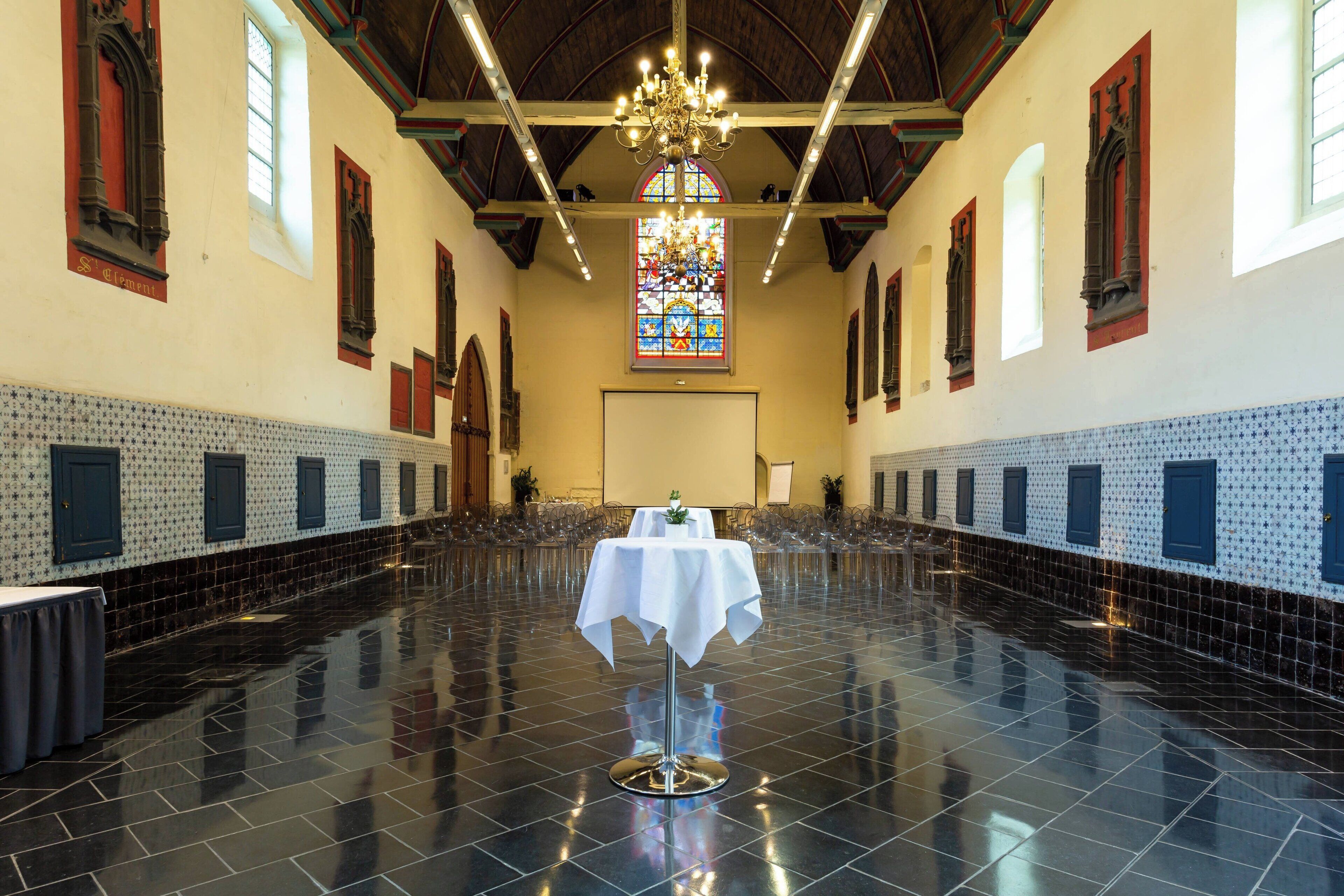 A spacious hall with a polished floor, a central table with a white tablecloth, and a large stained glass window.