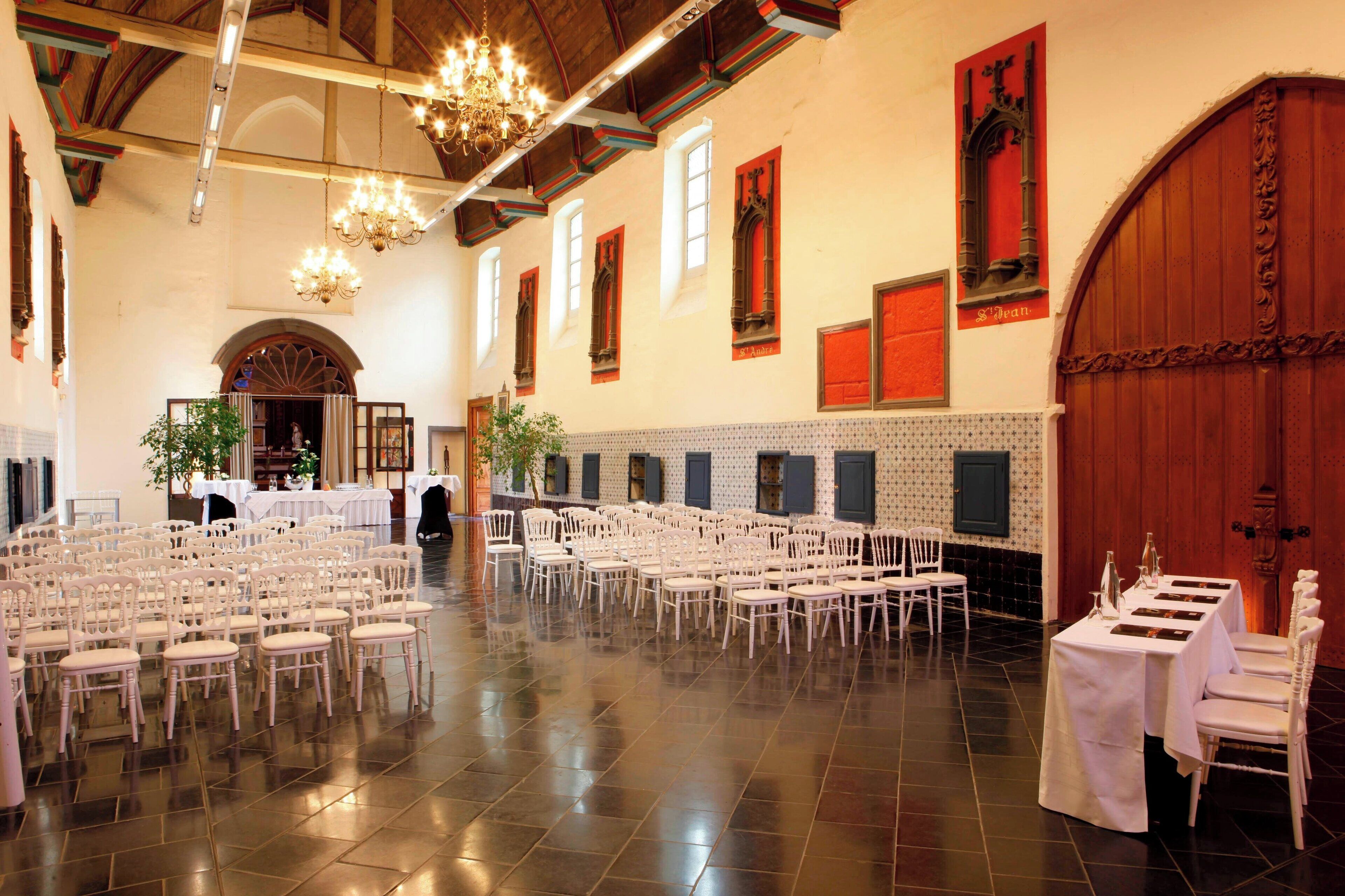 A spacious hall with rows of white chairs and tables, a chandelier, and a large wooden door.
