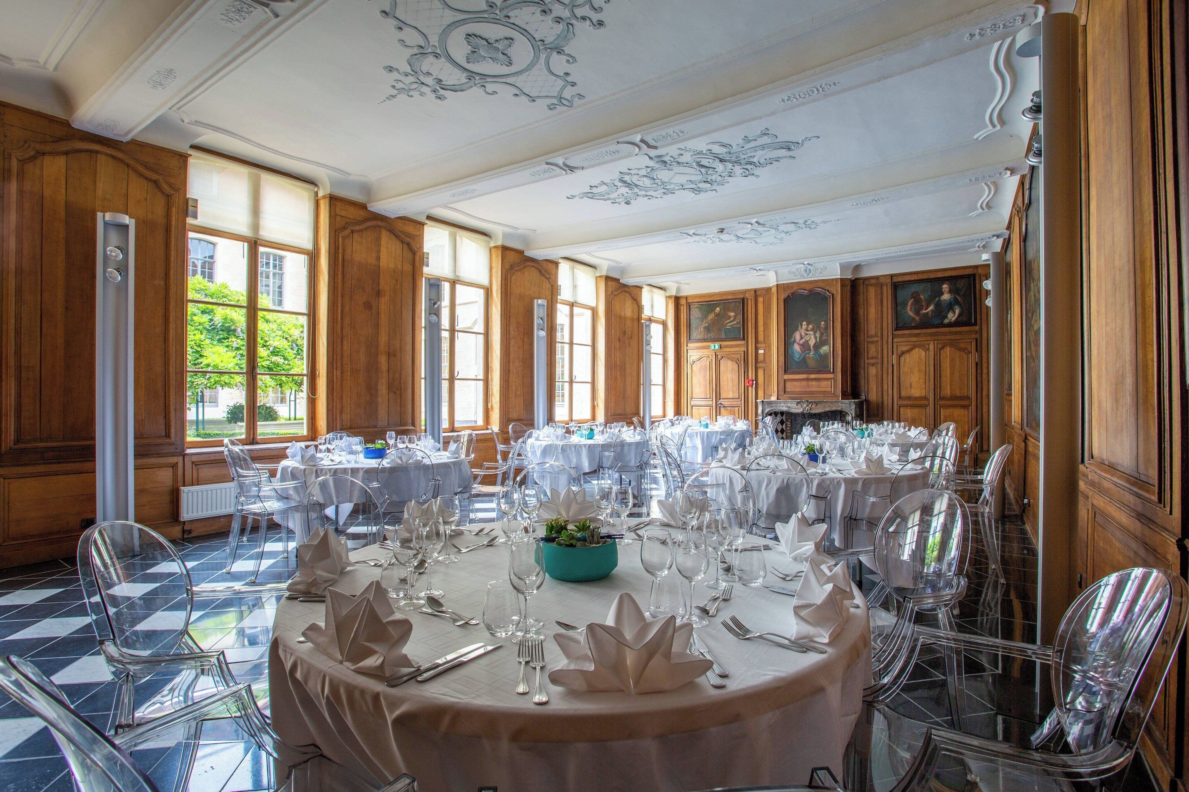 A dining area with round tables set for a formal event, featuring clear chairs and white tablecloths.