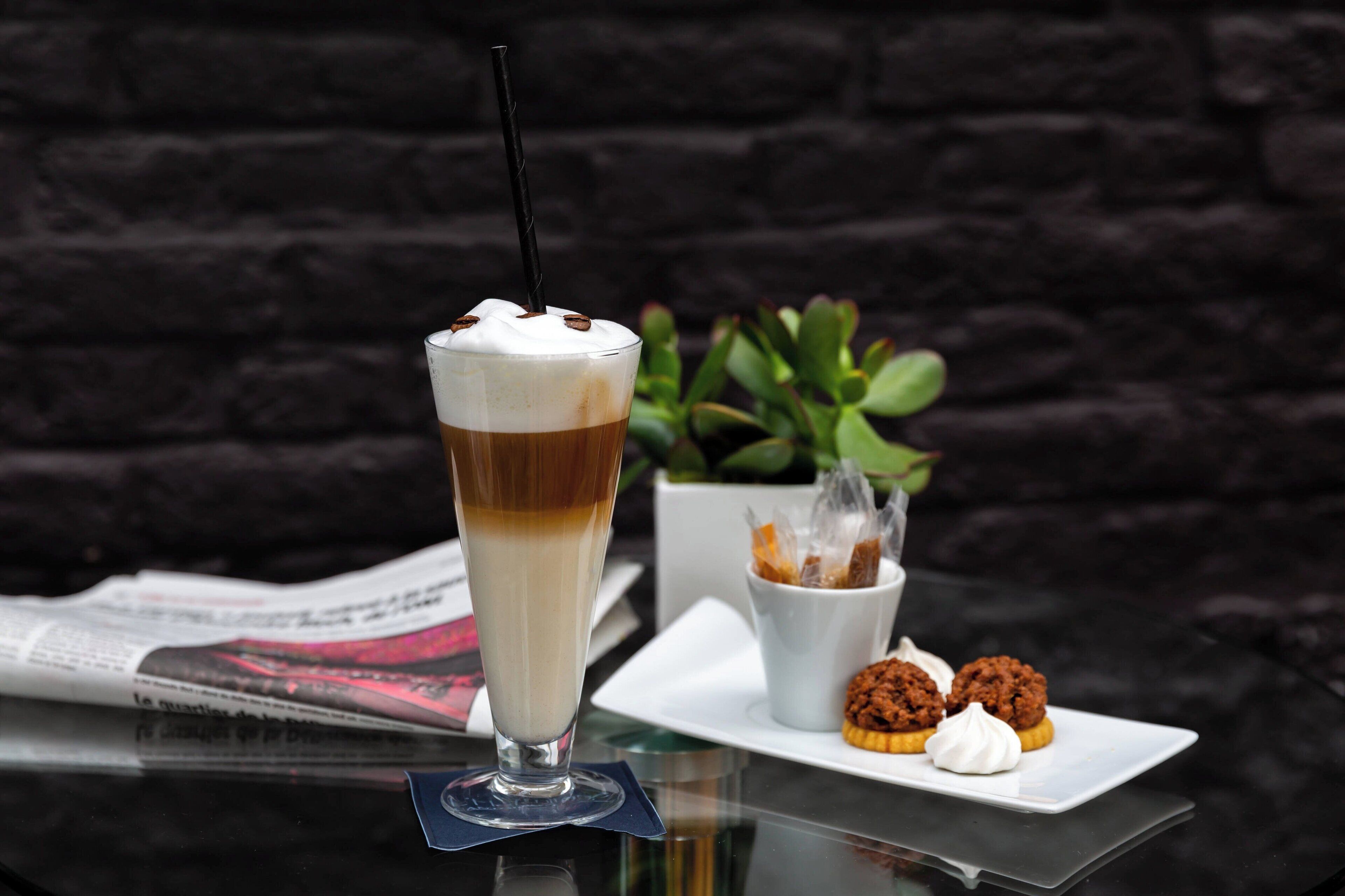 Een hoog glas met een gelaagde drank, een klein wit kopje met een lekkernij, en een bord met koekjes en een cupcake, alles op een glazen tafel, met een krant en een plantje op de achtergrond.