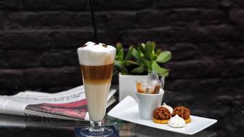 Een hoog glas met een gelaagde drank, een klein wit kopje met een lekkernij, en een bord met koekjes en een cupcake, alles op een glazen tafel, met een krant en een plantje op de achtergrond.
