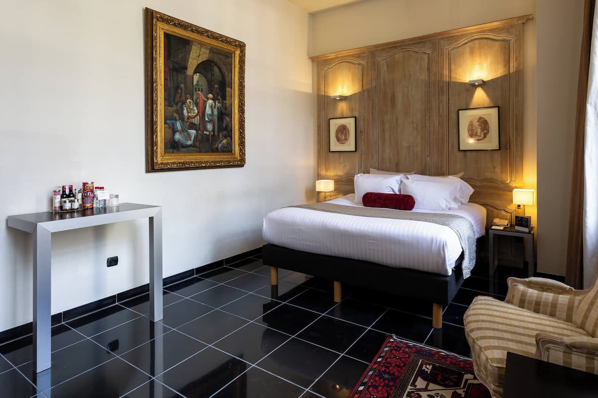 Une chambre d’hôtel avec un lit, des tables de chevet, une chaise, un tableau et un sol carrelé.