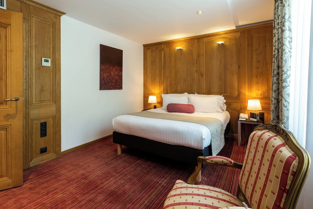 Une chambre d’hôtel avec un grand lit, une armoire en bois et un sol recouvert d’une moquette rouge.
