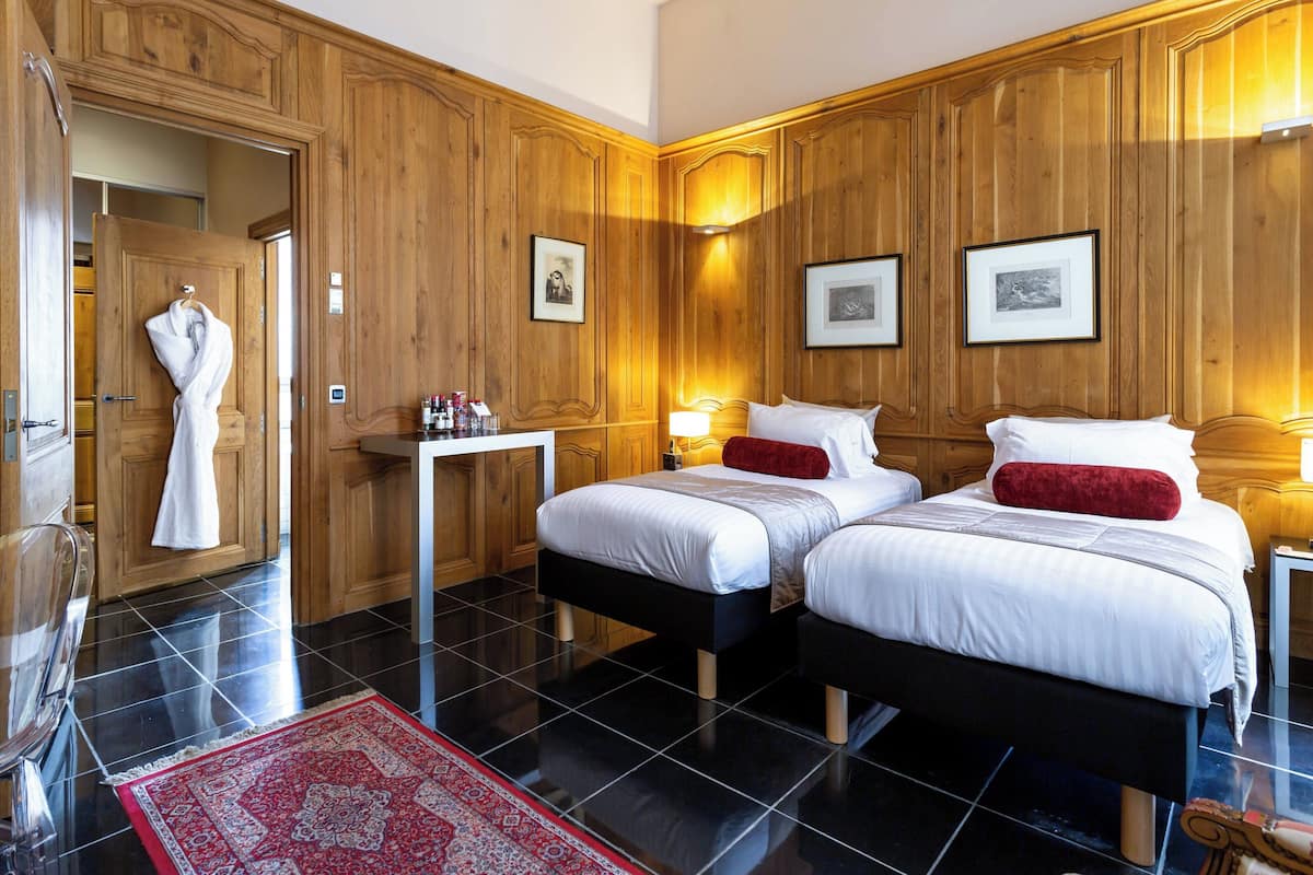 Une chambre d’hôtel avec deux lits, des boiseries, un tapis à motifs rouges et un peignoir accroché à la porte.