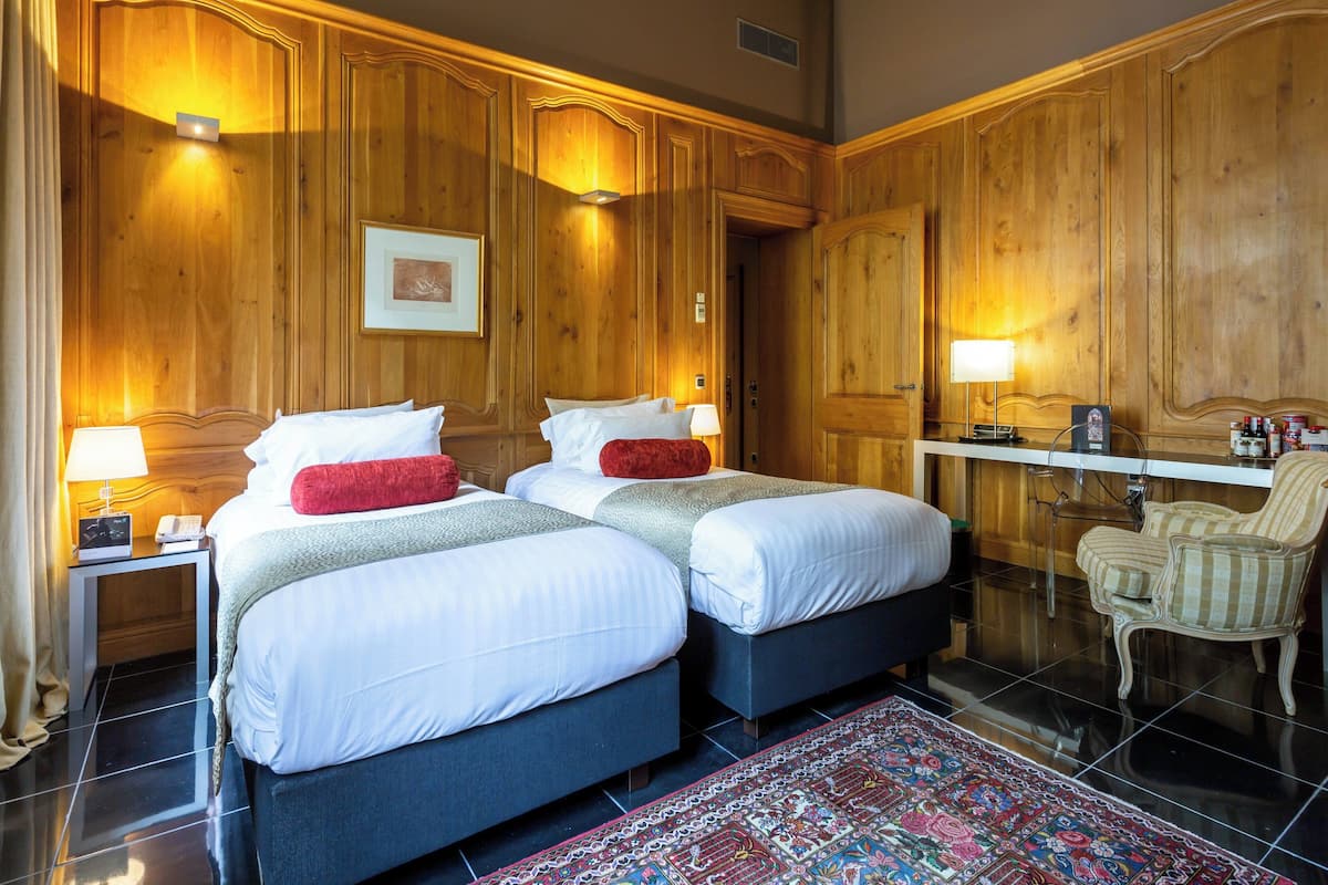 Une chambre d’hôtel avec deux lits, des boiseries, un bureau avec une lampe, une chaise et un tapis à motifs.