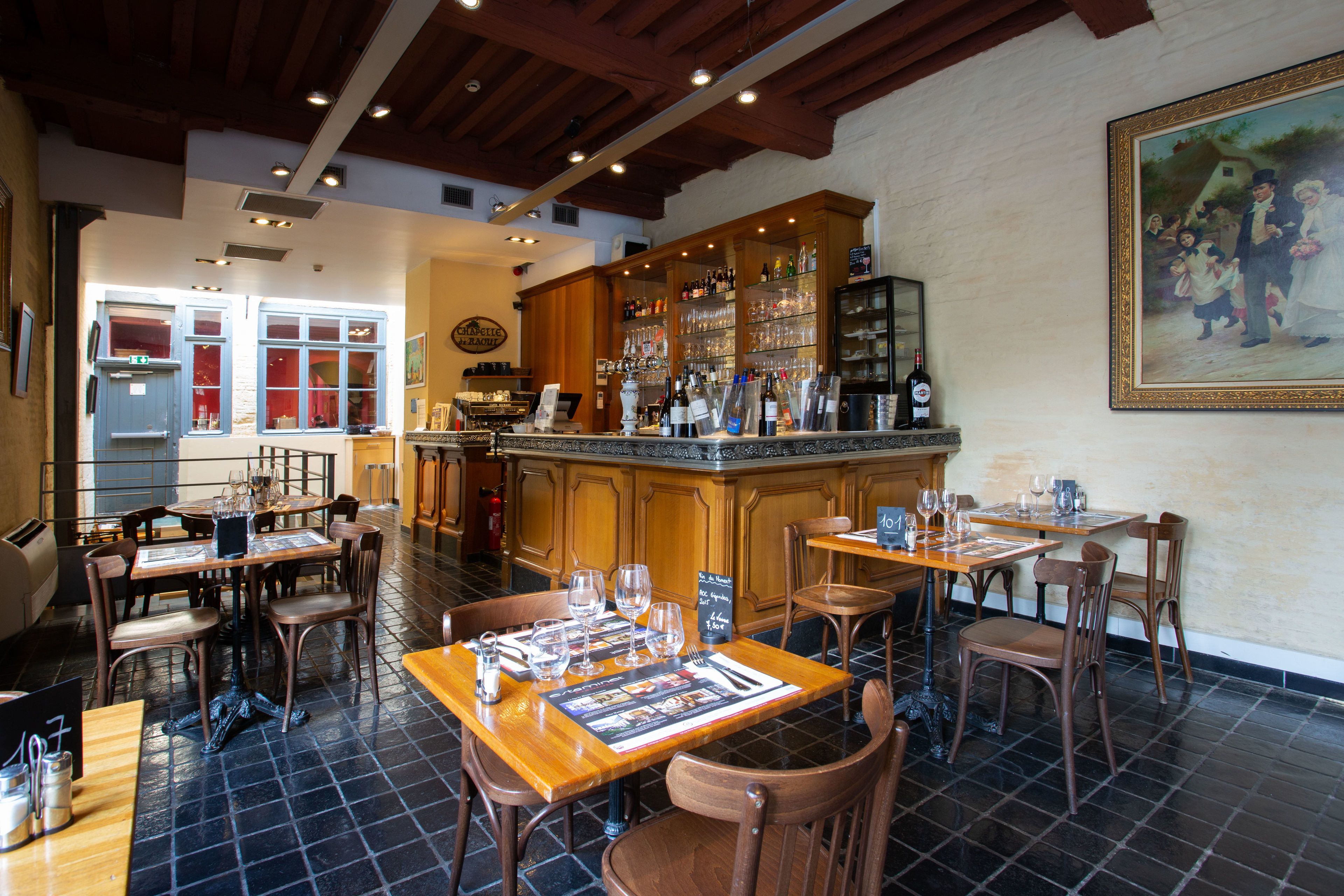 Un restaurant avec des tables et des chaises en bois, un coin bar avec des bouteilles exposées et un grand tableau accroché au mur.