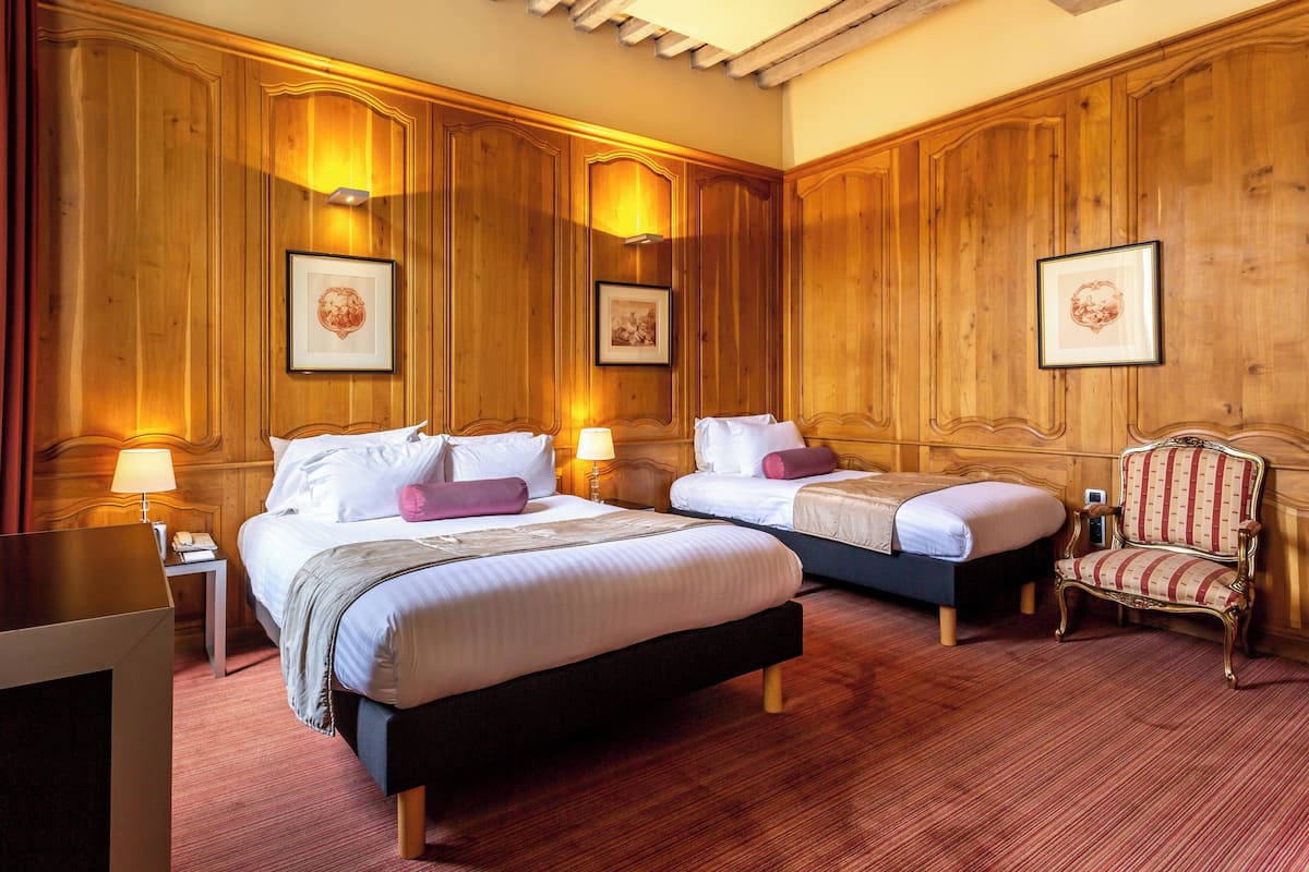 Une chambre d’hôtel avec deux lits, des boiseries, des œuvres d’art encadrées et un fauteuil classique.