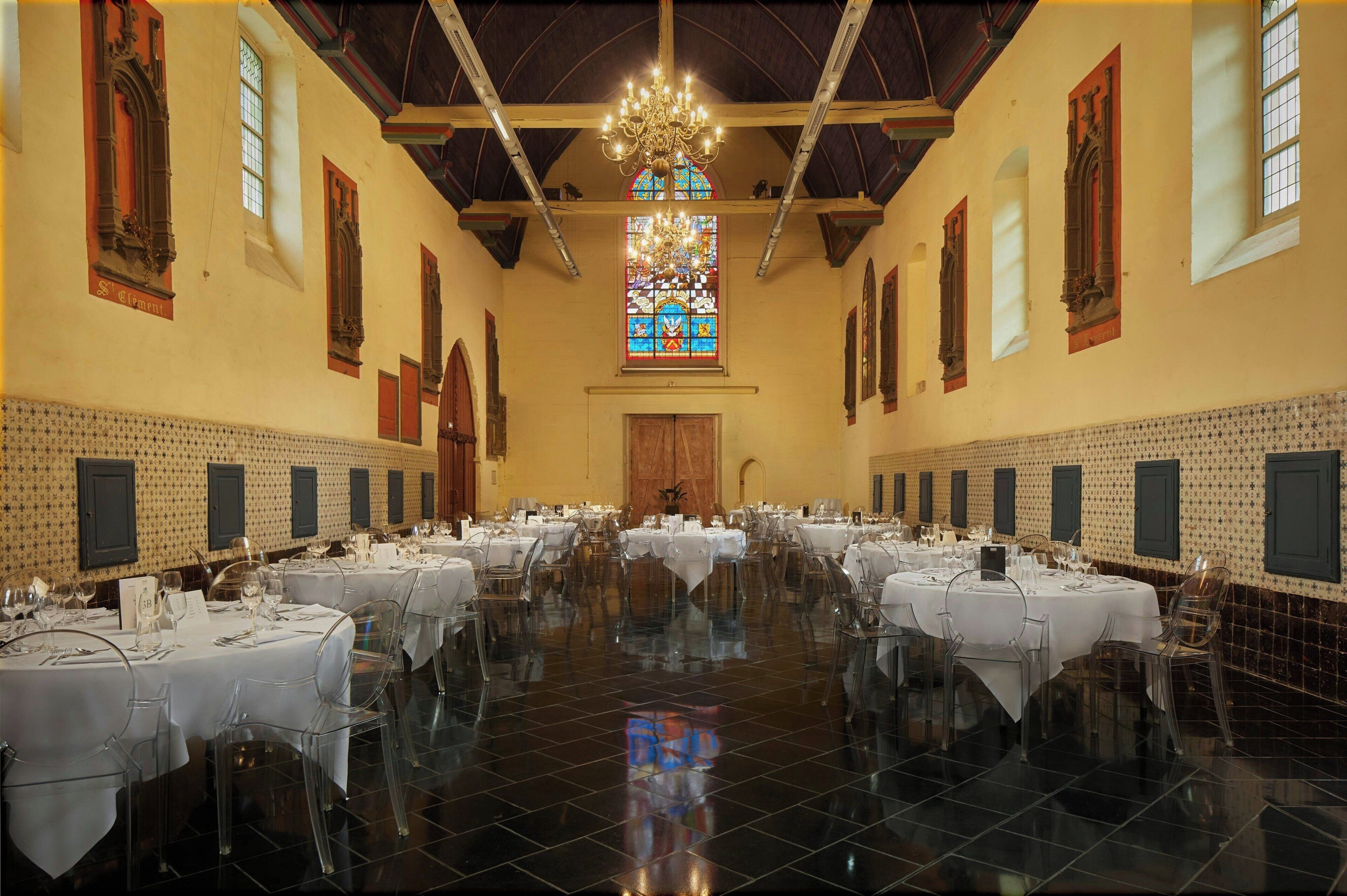 Une salle de banquet avec de nombreuses tables dressées pour le repas, une grande fenêtre à vitraux et un lustre.