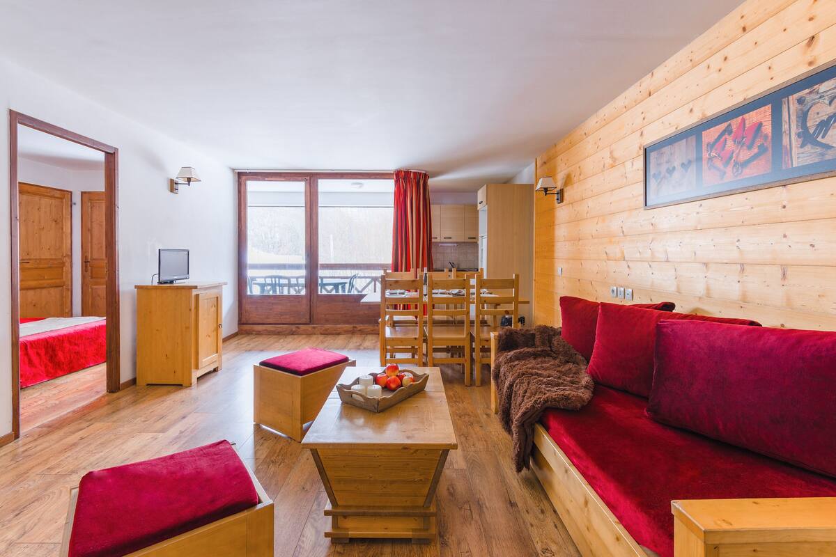 A cozy room with a wooden floor, a red sofa, a wooden table with fruit, and a view of a snowy landscape through large windows.