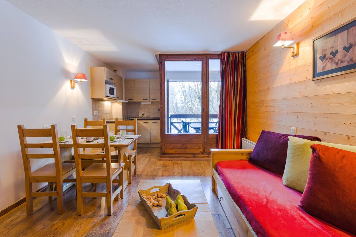 A cozy room with a wooden floor, a red sofa, a wooden dining table with chairs, and a kitchen area visible through an open door.