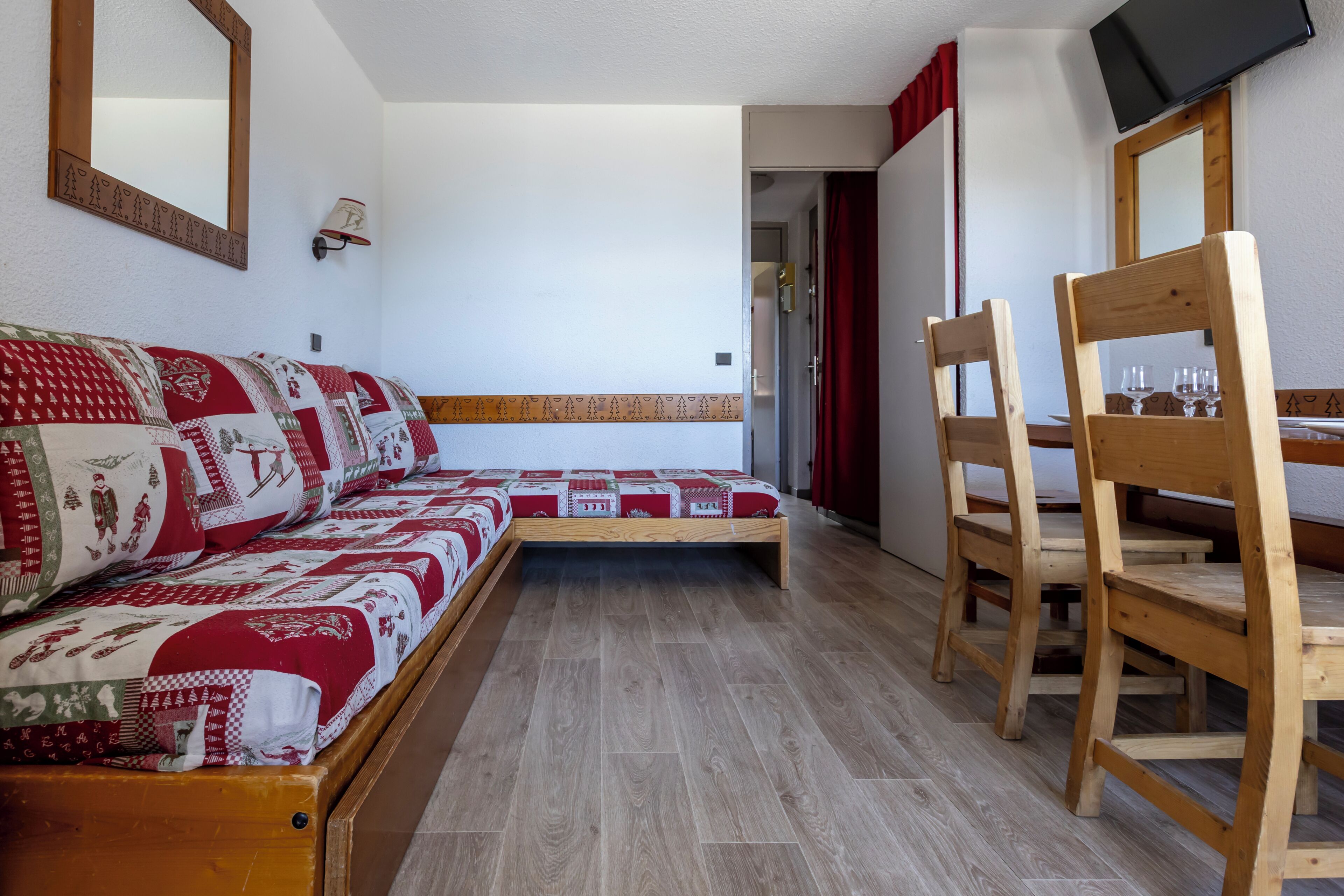 Una habitación con una cama y una silla de madera, una mesa con vasos, una lámpara de pared y un televisor.