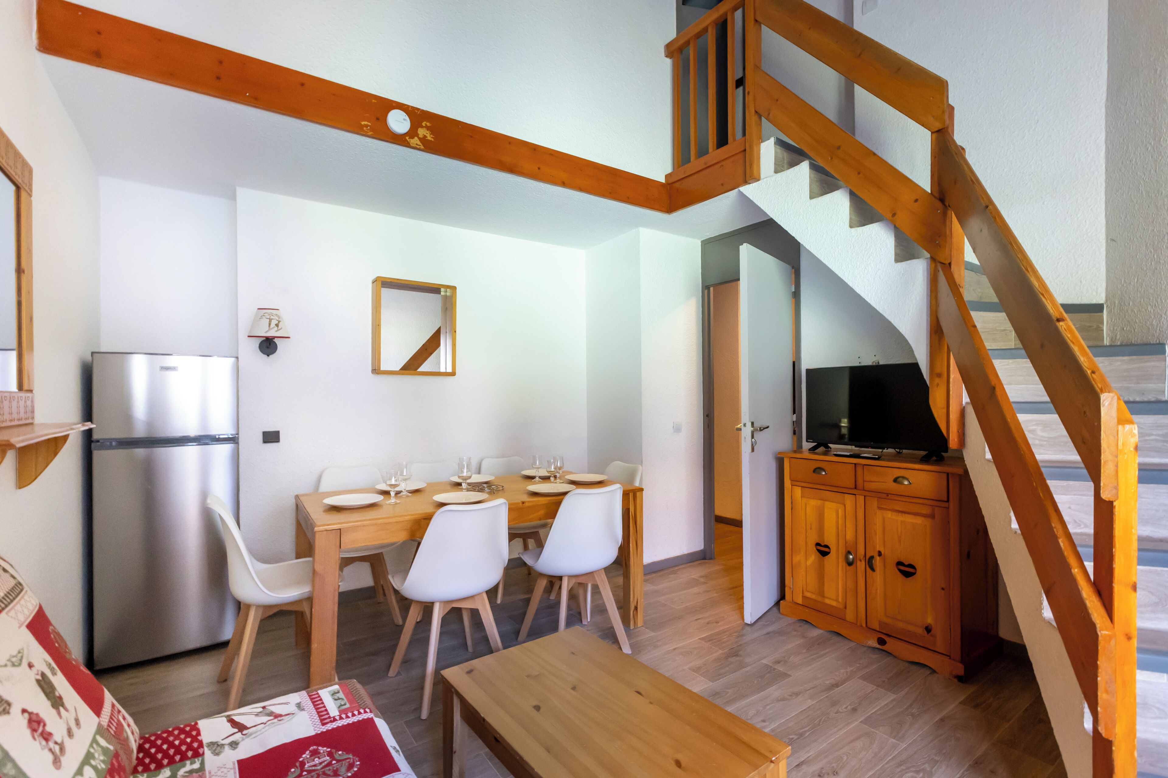 Un coin repas avec une table et des chaises en bois, un réfrigérateur, un téléviseur posé sur un meuble en bois, et un escalier avec des rampes en bois.