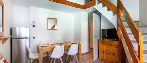 A dining area with a wooden table and chairs, a refrigerator, a television on a wooden cabinet, and a staircase with wooden railings.