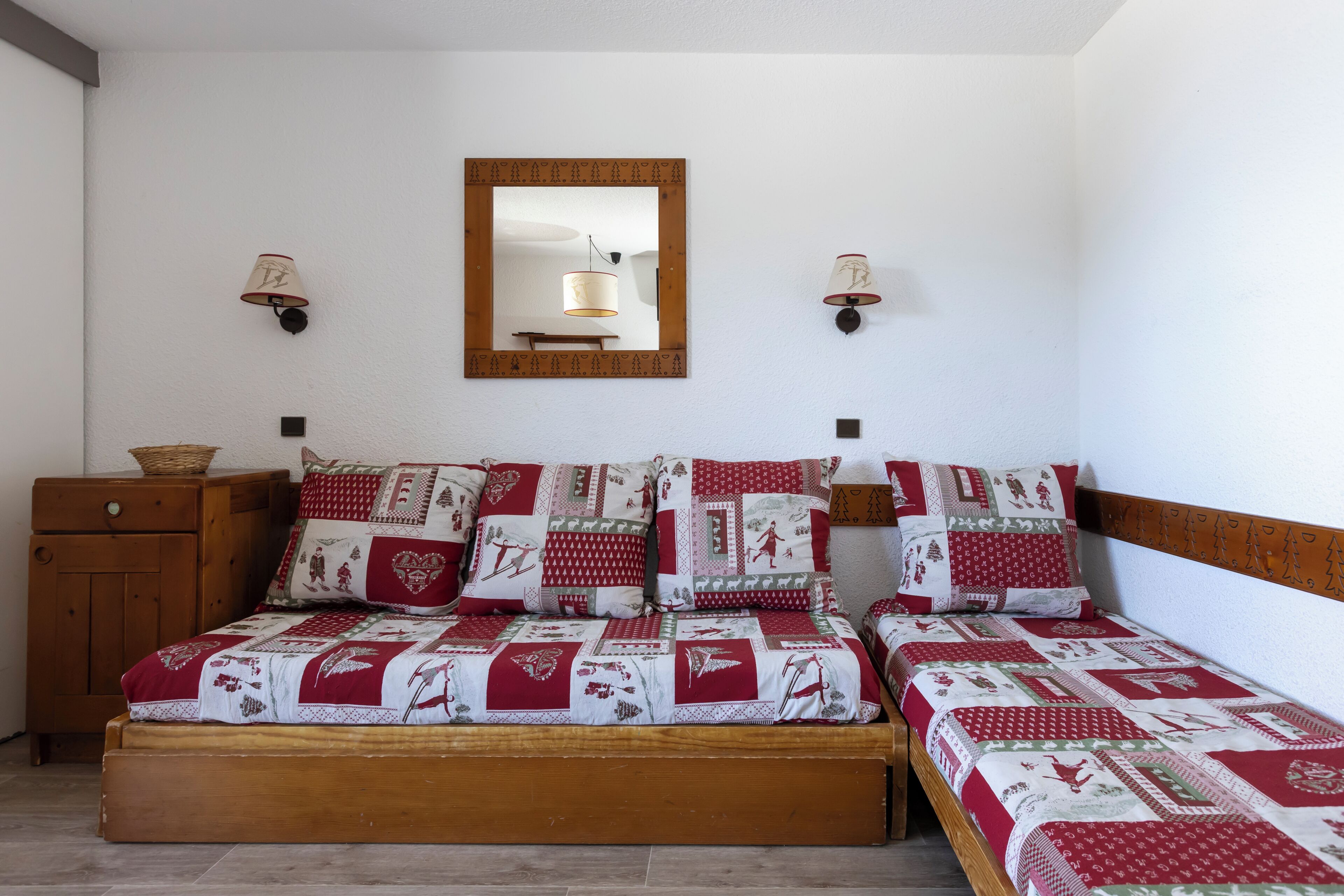A double bed with a red and white quilt, two wall-mounted lamps, a wooden nightstand, and a mirror on the wall.