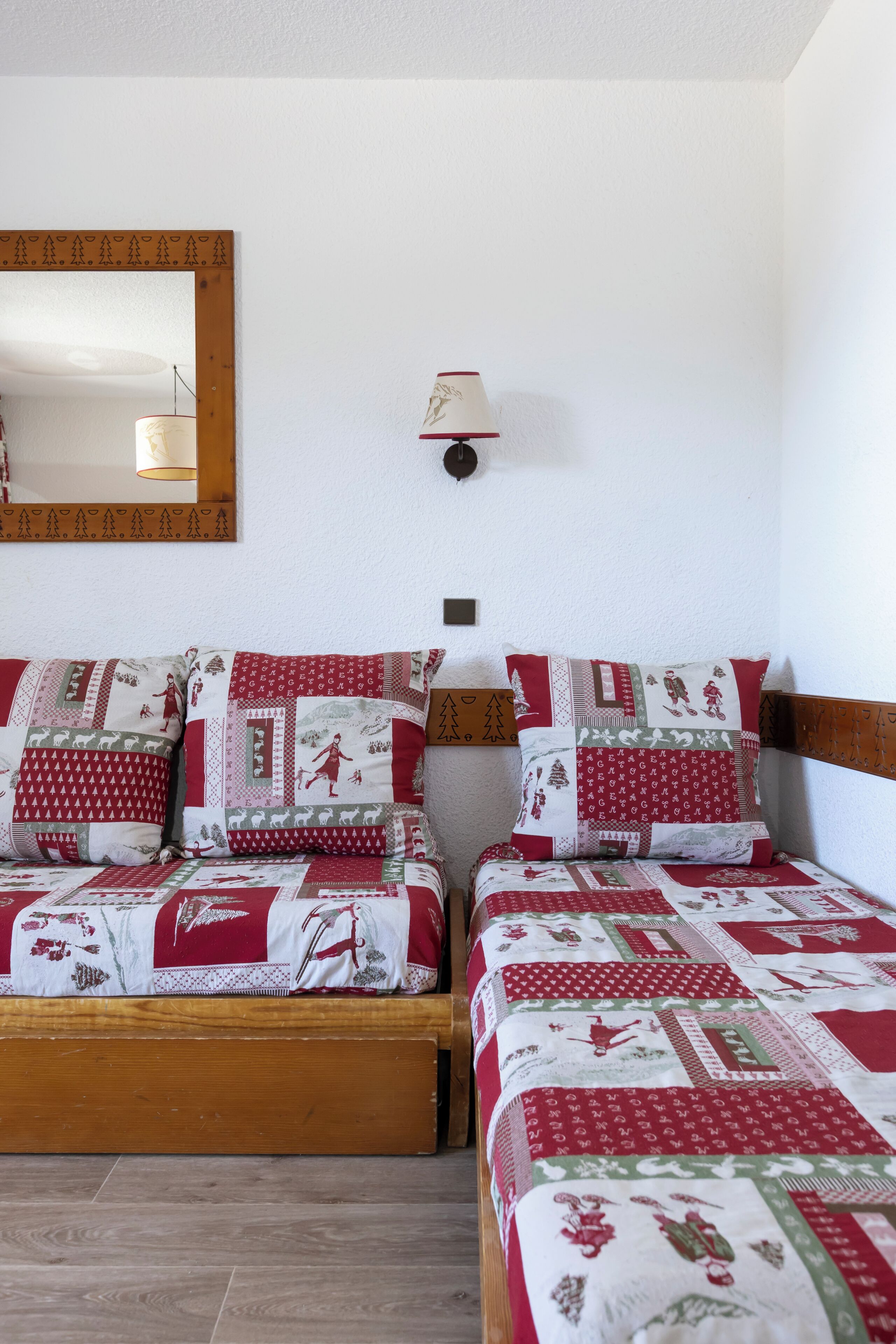 Un dormitorio con dos camas de madera, cada una con una colcha roja y blanca. Hay un espejo en la pared y una lámpara de noche.