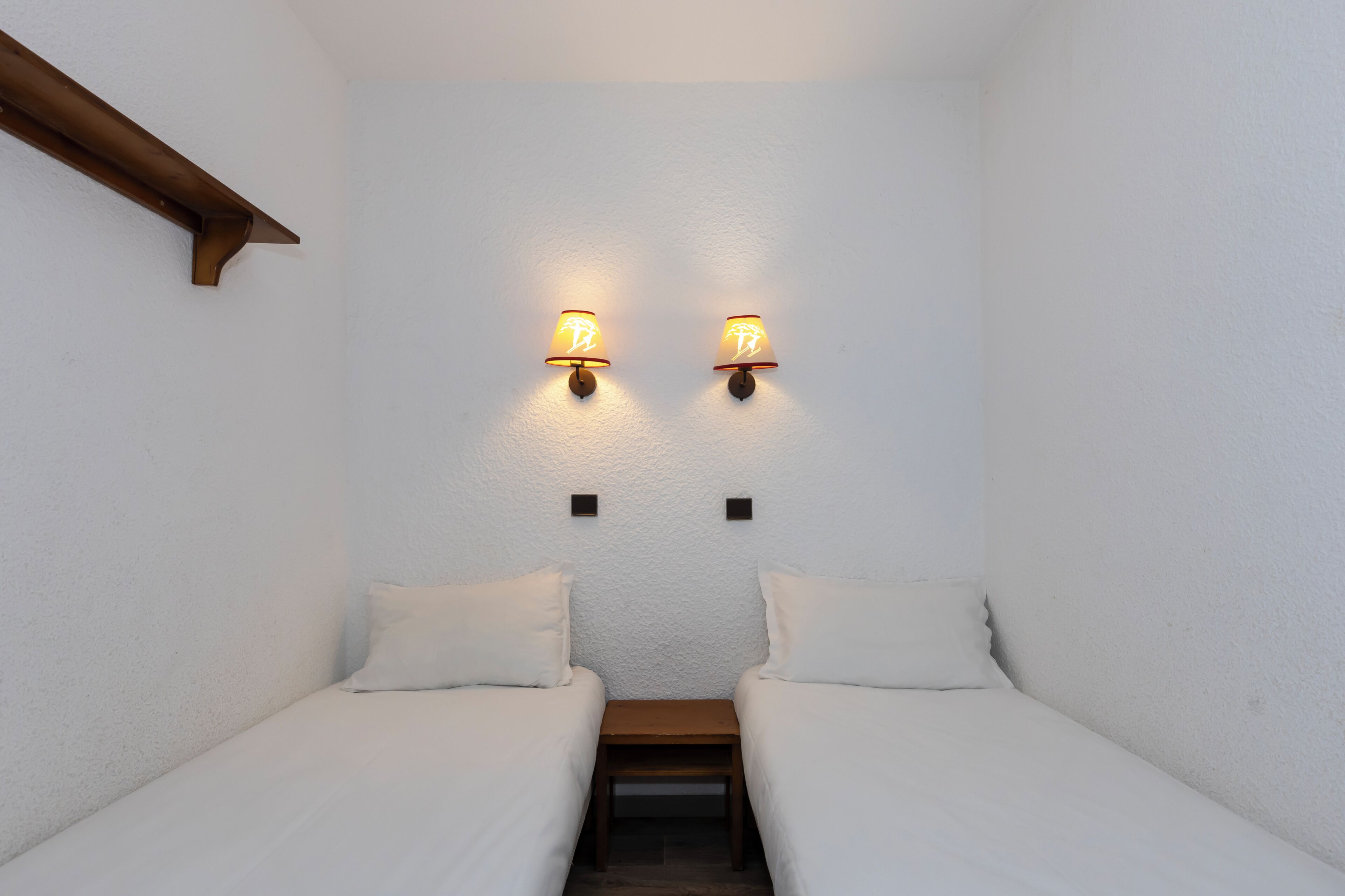 A small, cozy room with two single beds, a wooden bench, and wall-mounted lamps.