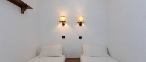 A small, cozy room with two single beds, a wooden bench, and wall-mounted lamps.