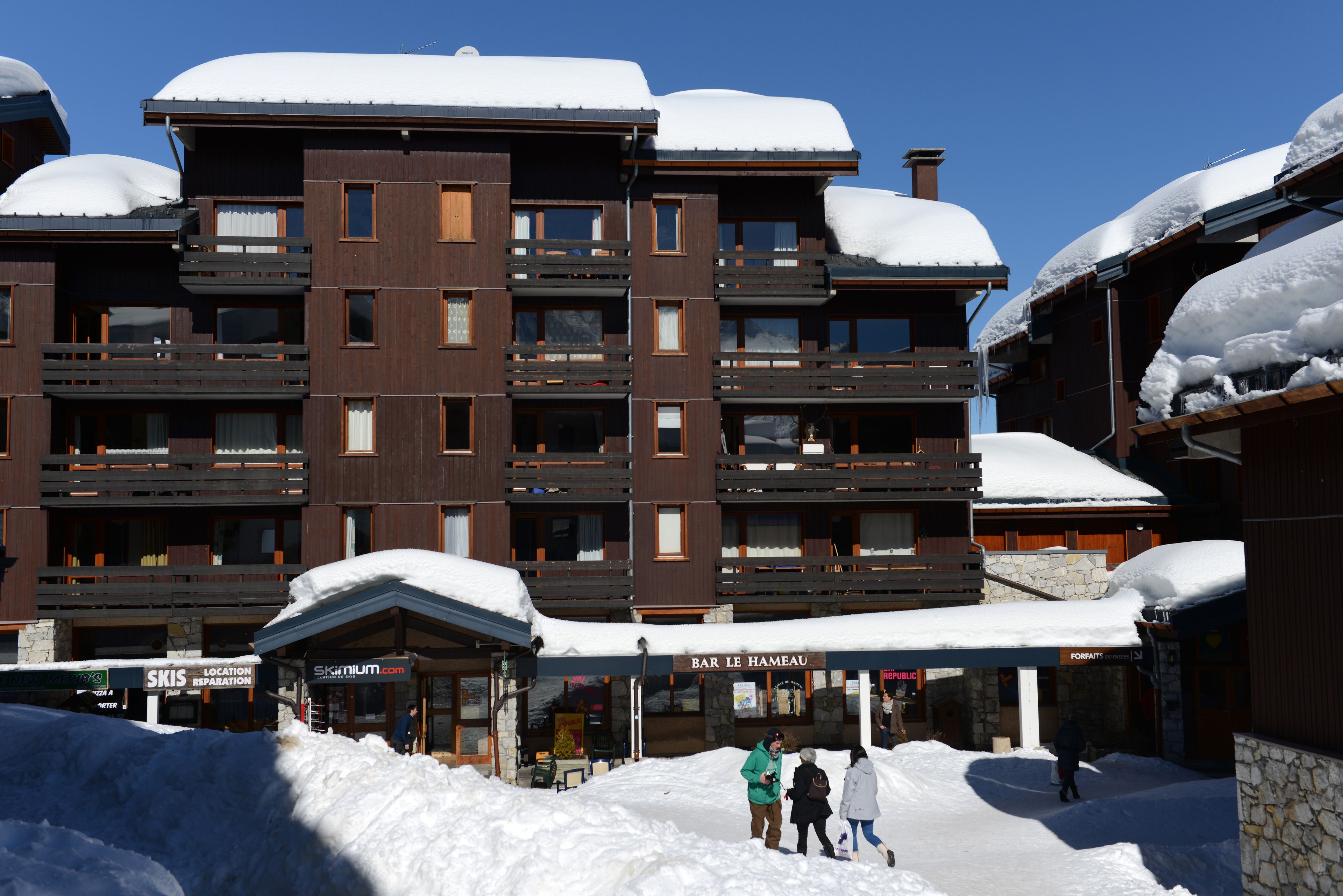 Ein mehrstöckiges Gebäude mit Balkonen, einem Schild für einen Skiort und einer Bar namens "Bar Le Hameau".
