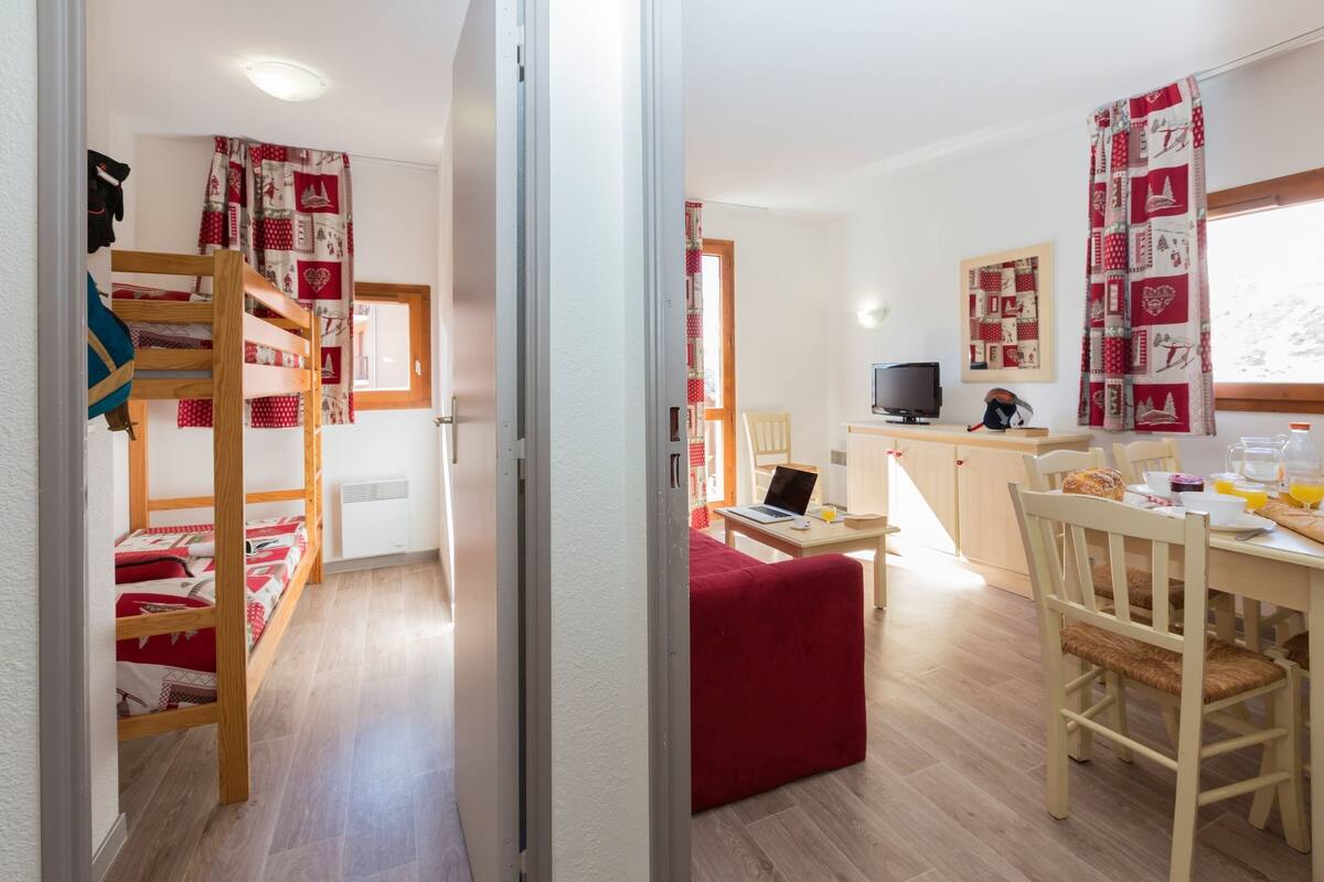 A room with a bunk bed, a desk with a laptop, a dining table with chairs, and a television.