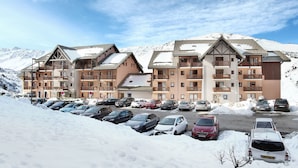 A multi-story hotel with a snow-covered parking lot and mountainous backdrop. - Résidence Odalys Les Lumières de Neige (Valmeinier)