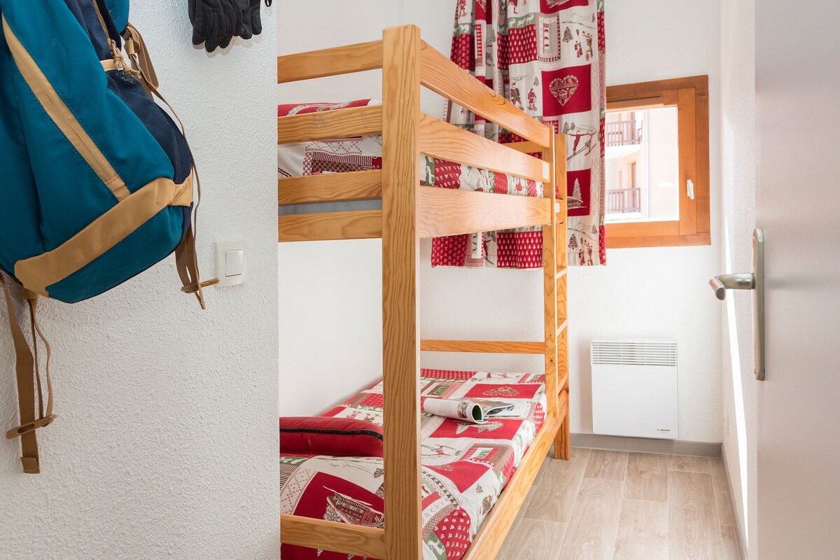 A bunk bed with a red and white patterned bedspread, a wooden door, and a window with a view of a balcony.