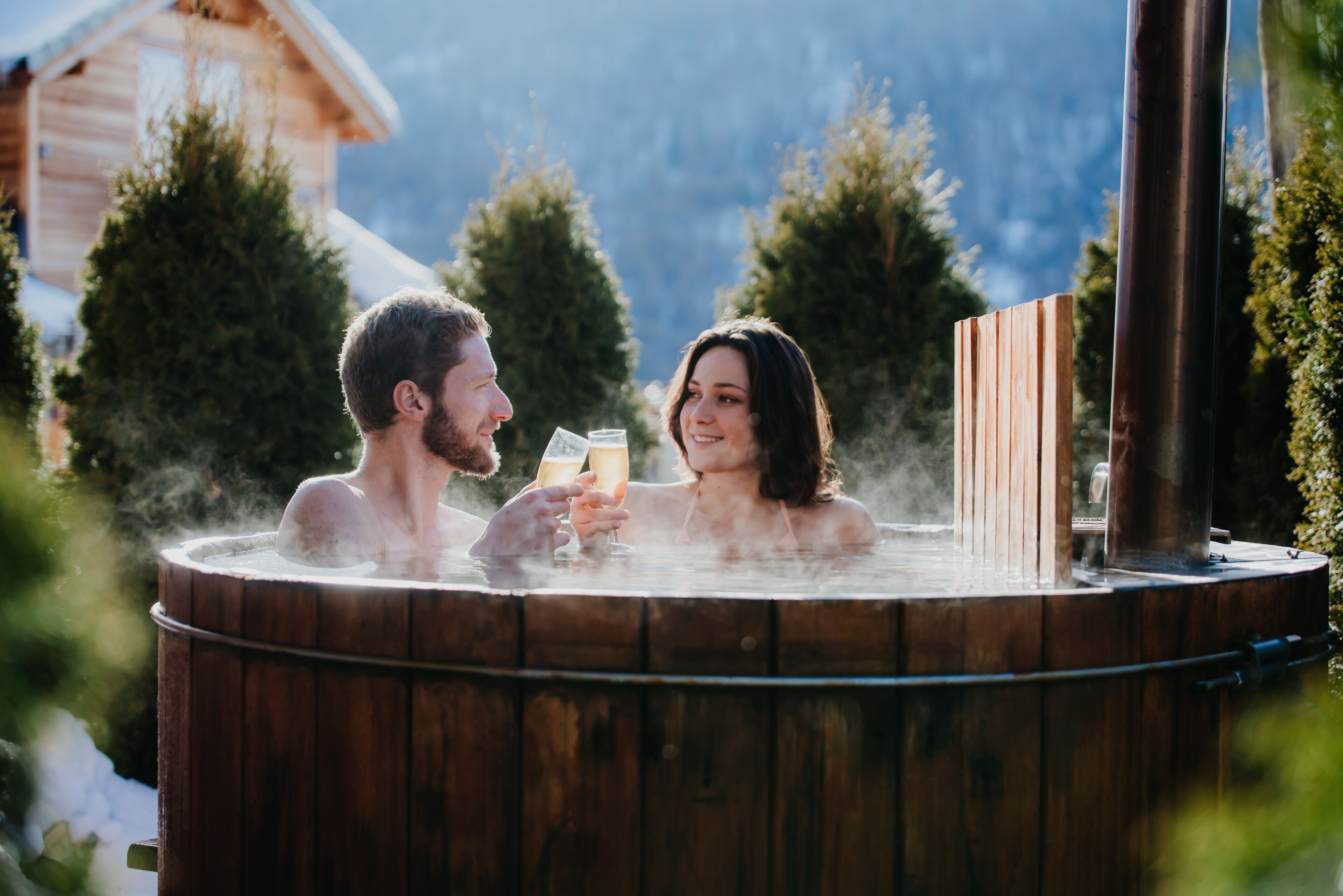 Una pareja en un jacuzzi con revestimiento de madera, rodeada de árboles y una montaña al fondo.
