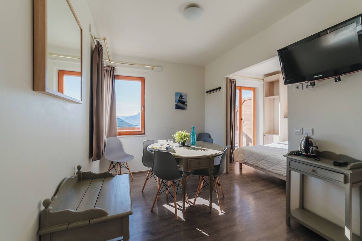 A room with a bed, a TV, a desk, a dining table with chairs, and a window with curtains.
