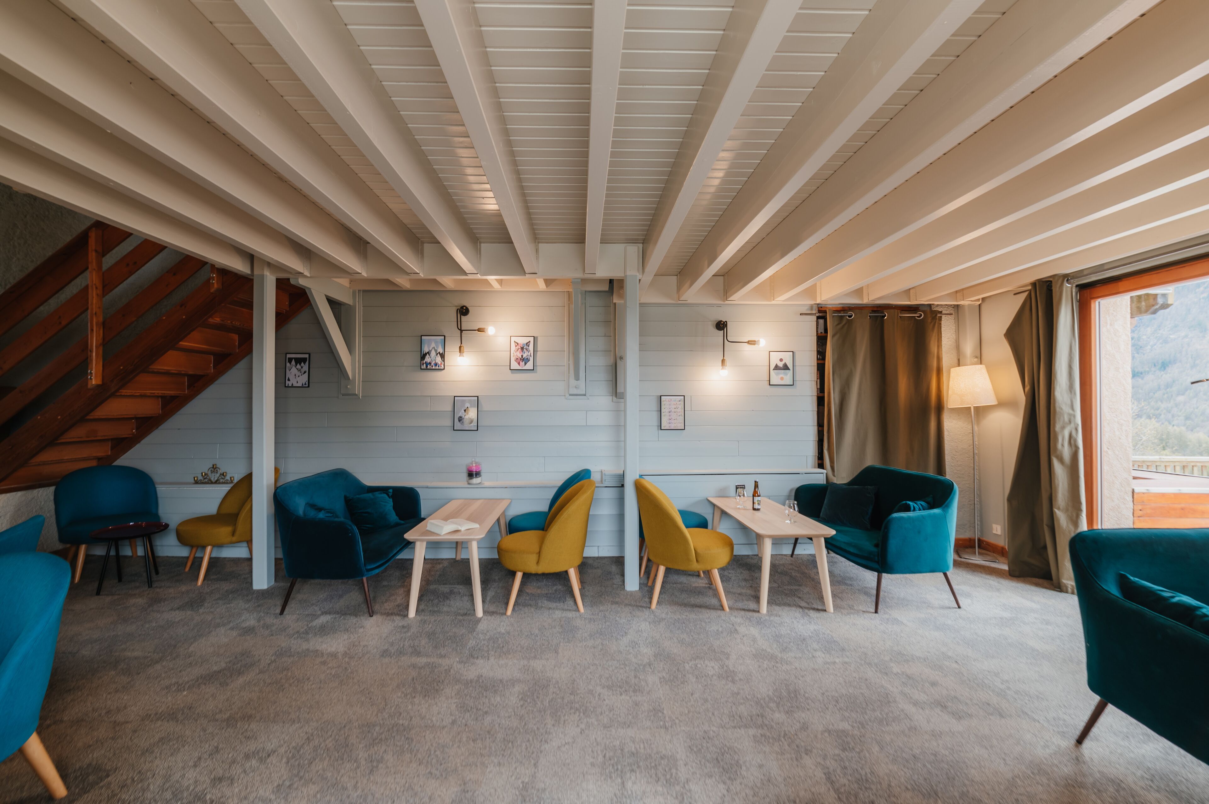 Un interior moderno con una escalera, vigas de madera y una mezcla de sillas azules y amarillas alrededor de una mesa central.