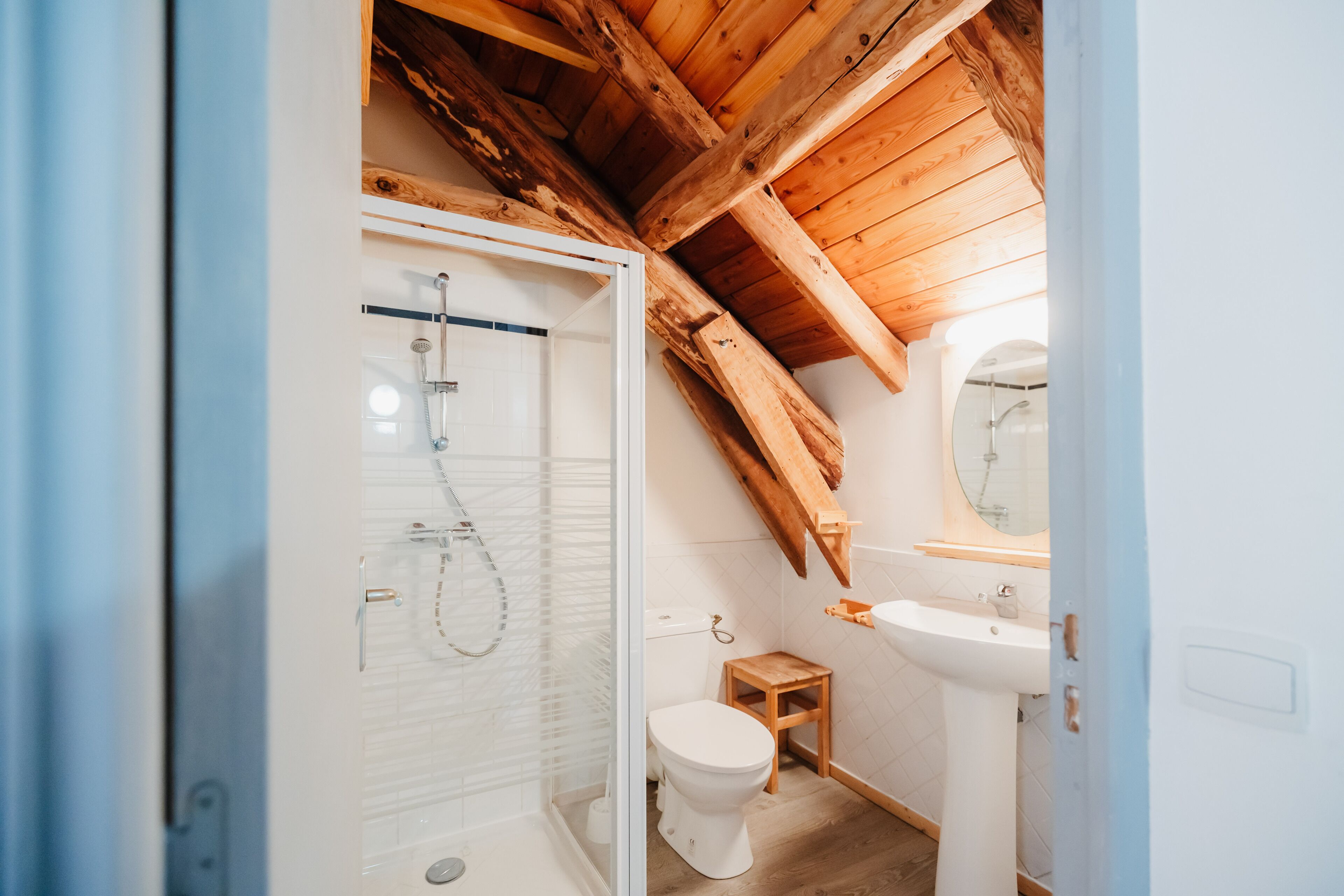 Ein kleines, rustikales Badezimmer mit Holzbalkendecke, einer Duschkabine aus Glas, Toilette und Waschbecken.