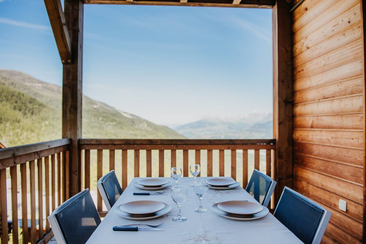 A wooden deck with a table set for two, overlooking a mountainous landscape.