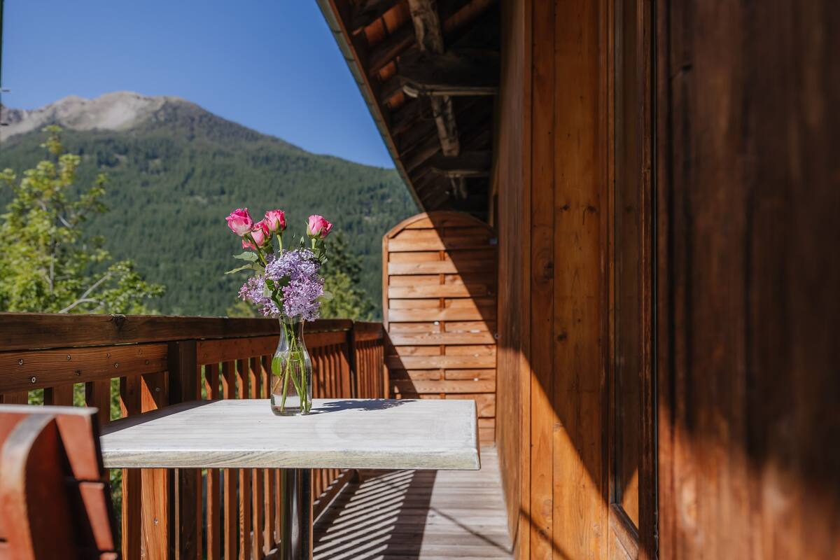 A wooden balcony with a table and chairs, a vase with flowers, and a mountain view in the background.