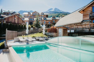 Un espace piscine avec des chaises longues, un bâtiment en bois doté de balcons et une vue sur les montagnes en arrière-plan.
