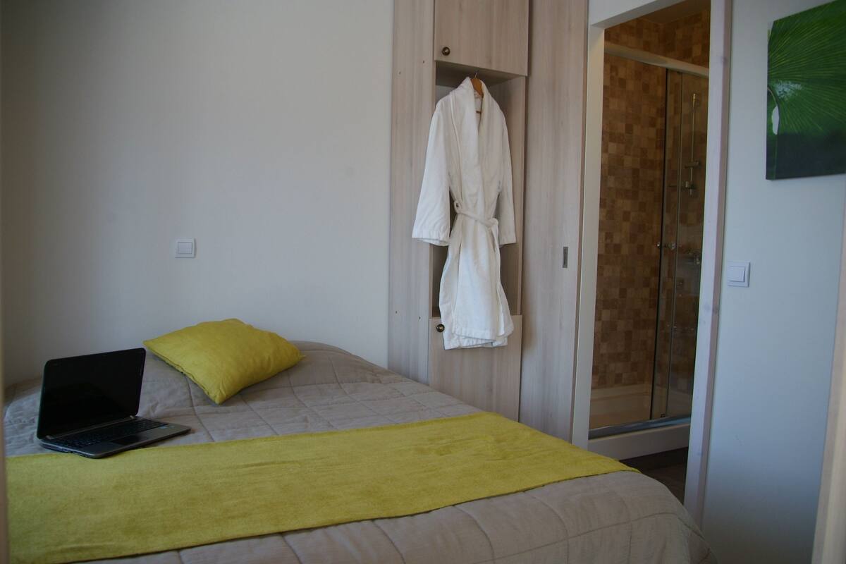 A bedroom with a bed, a laptop, a yellow pillow, a white robe hanging on a door, and a shower area.
