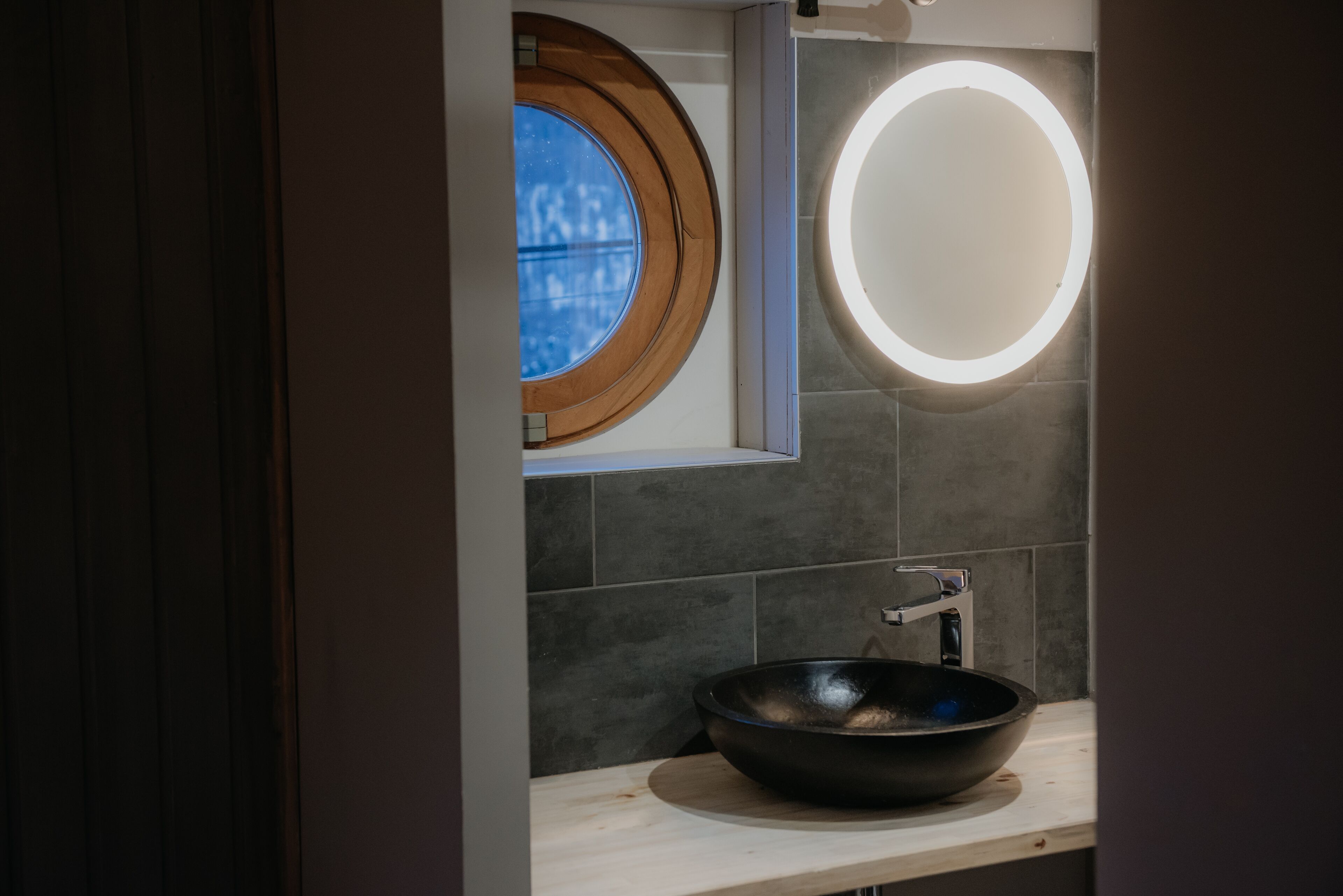 Un baño moderno con un espejo redondo, una ventana con marco de madera y un lavabo negro.